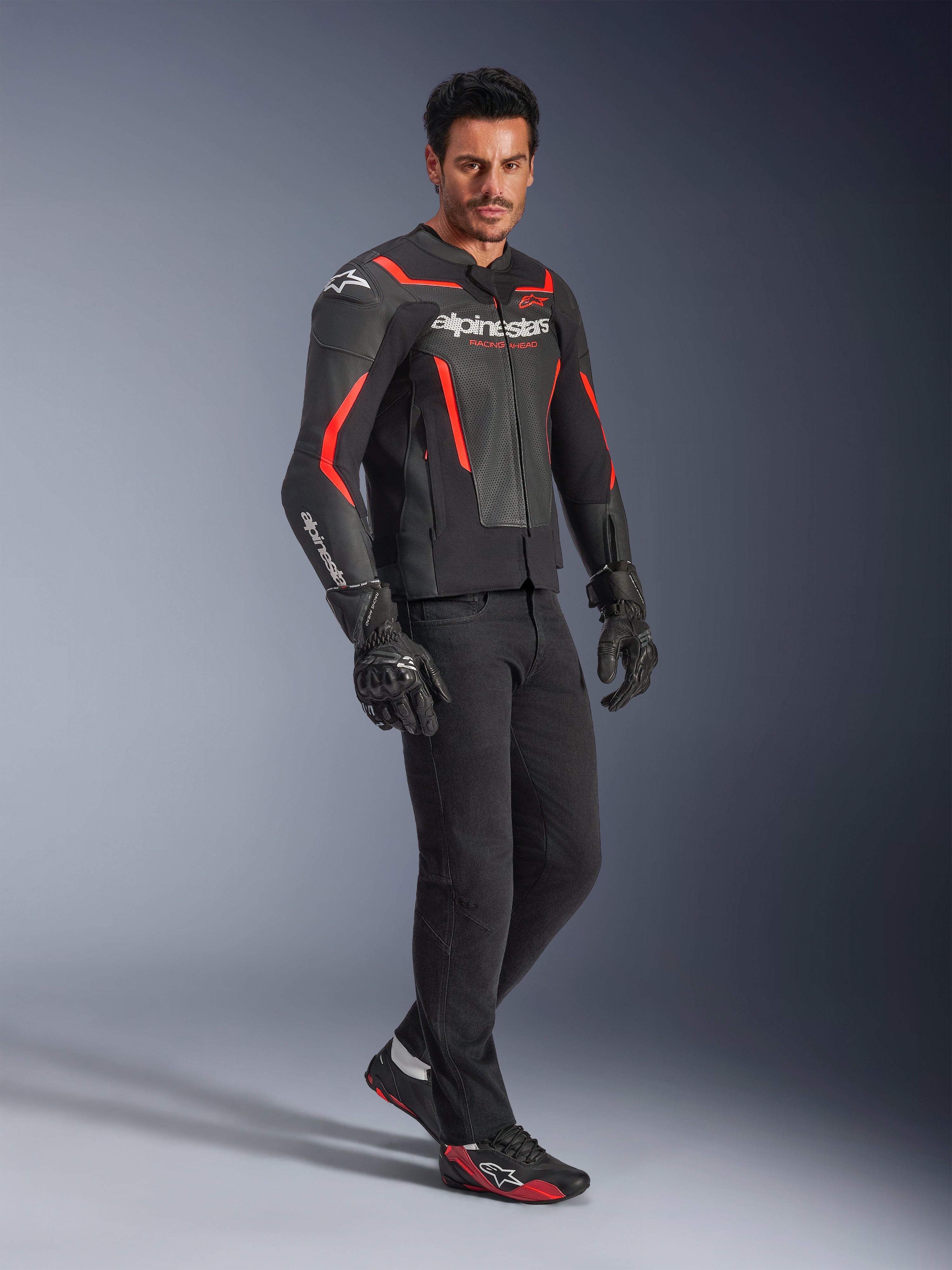 Modèle portant le blouson en cuir Alpinestars GP Force V2 Air en noir et rouge fluo, associé à un pantalon de moto noir, des gants et des baskets assorties, avec des panneaux en cuir perforé et le logo Alpinestars blanc, équipement de moto sport