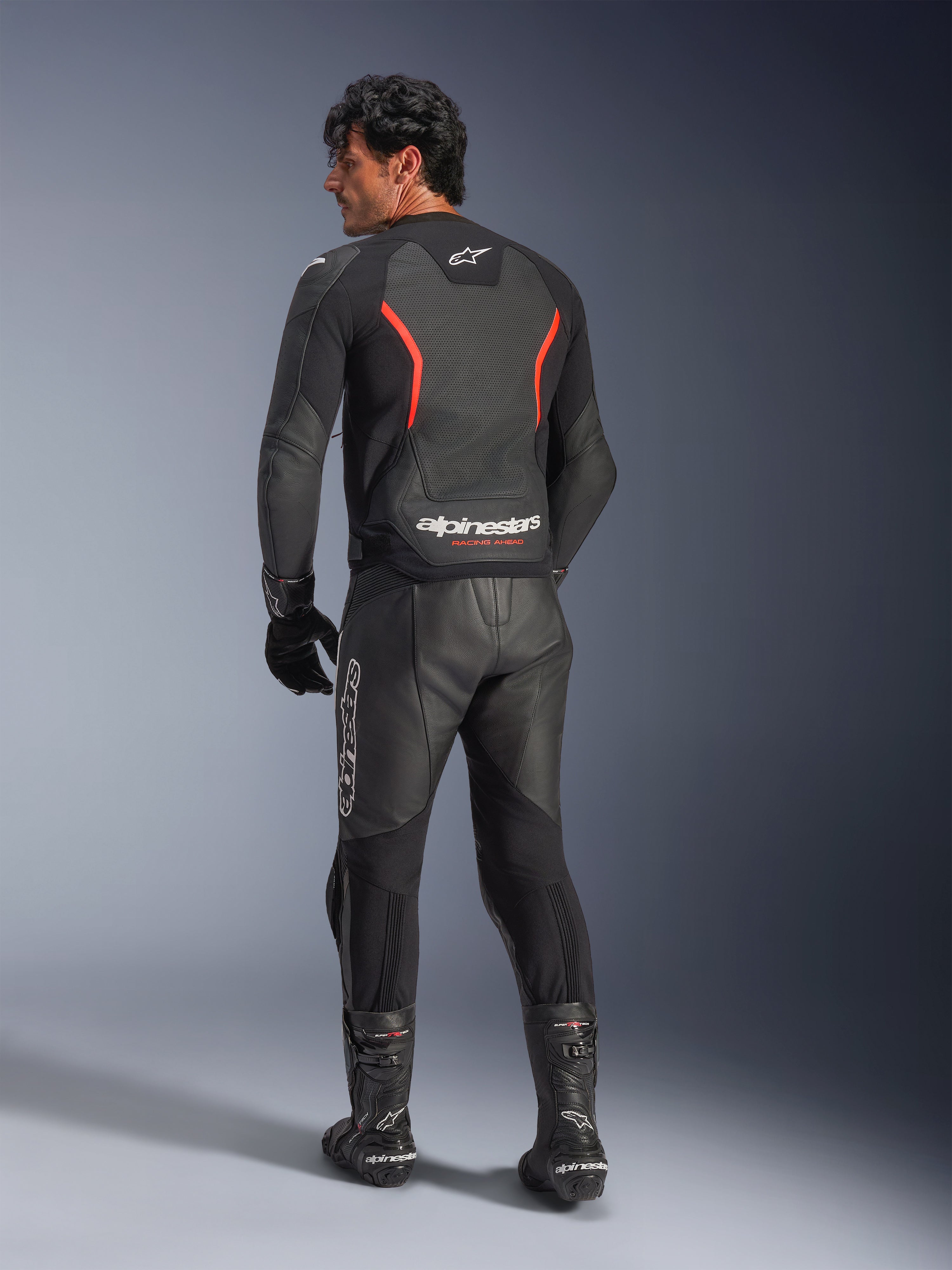 Vue arrière d'un mannequin portant le blouson en cuir Alpinestars GP Force V2 Air en Noir Rouge Fluo, doté de panneaux en cuir perforés et d'un logo Alpinestars distinctif au bas du dos, associé à un pantalon et des bottes de moto en cuir noir assortis.