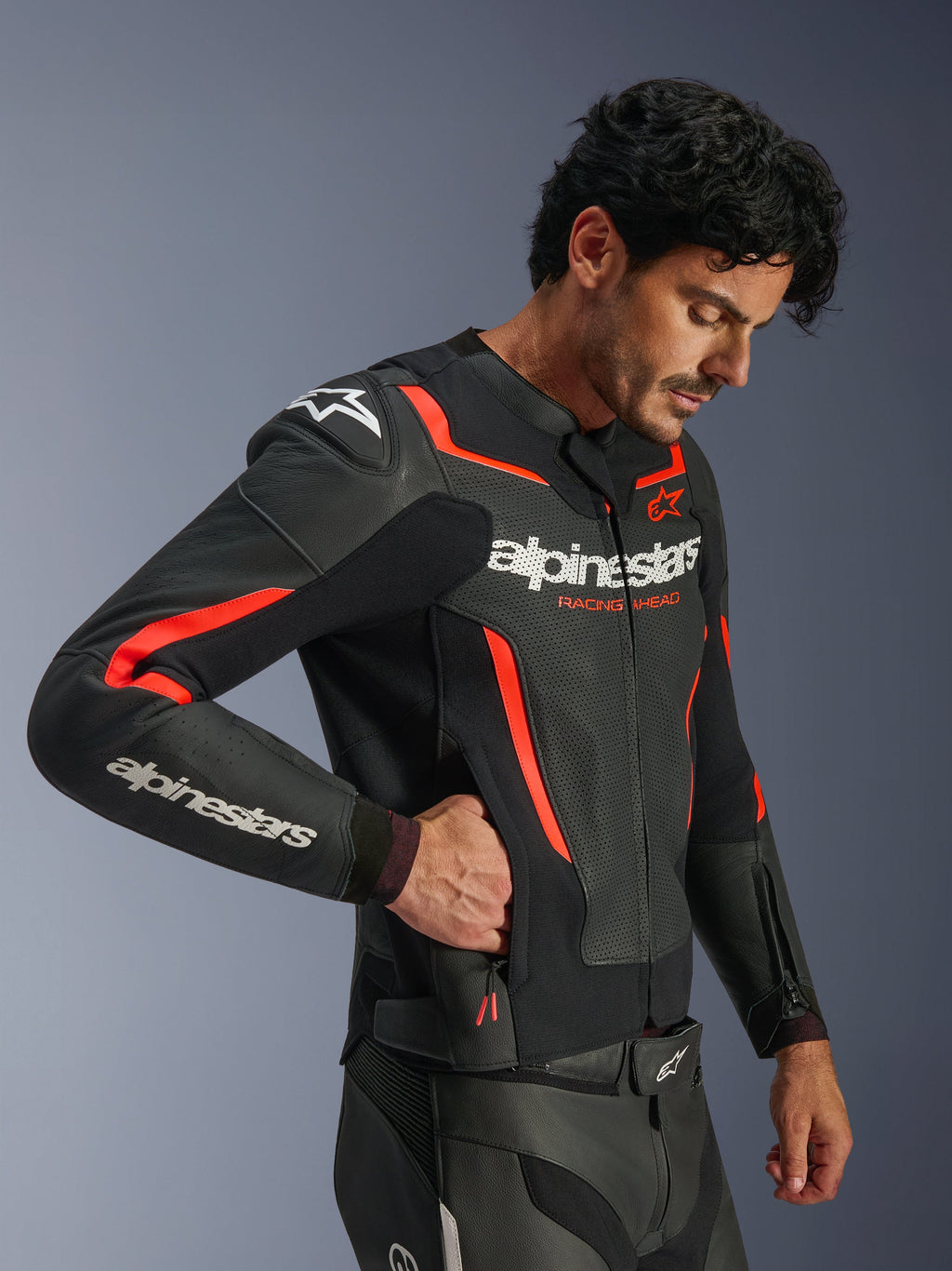 Blouson en cuir Alpinestars GP Force V2 Air, noir et rouge fluo, blouson de moto en cuir perforé, mannequin portant le blouson et mettant la main dans une poche latérale zippée, avec des accents rouge fluo et logos Alpinestars blancs, équipement de conduite sportive professionnel