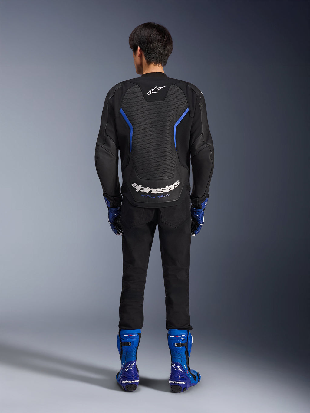 Blouson en cuir Alpinestars GP Force V2 Airflow, Noir Bleu Métallique, vue arrière montrant des panneaux en cuir perforé et un marquage blanc, porté avec des bottes de course bleues et un pantalon noir, équipement de course moto sportive