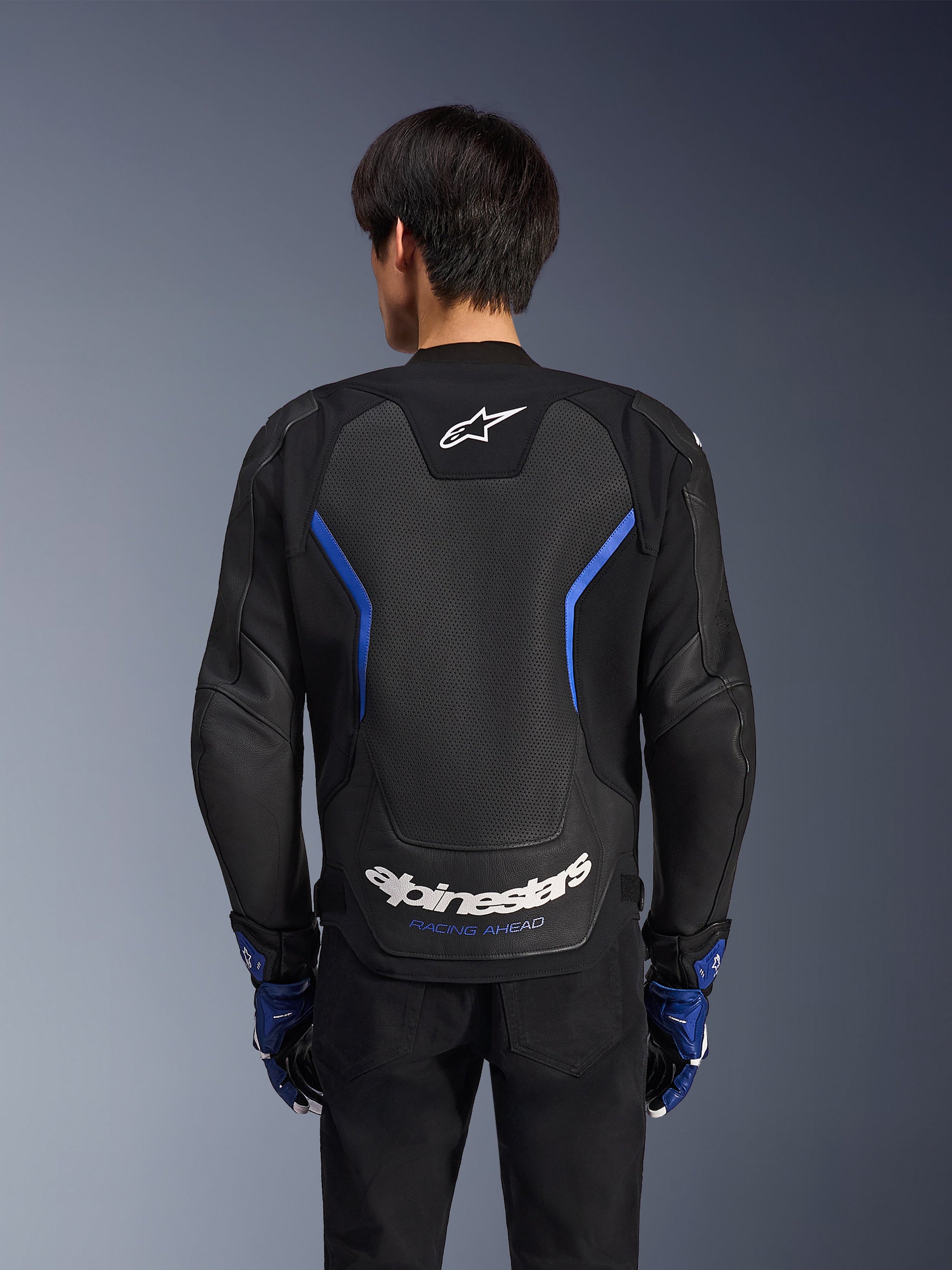 Blouson en cuir Alpinestars GP Force V2 Airflow, Noir Bleu Métallisé, vue arrière, doté d'un grand panneau perforé pour la ventilation, d'accents bleus et du logo Alpinestars blanc, porté avec des gants bleus assortis et un pantalon noir pour la conduite sportive.