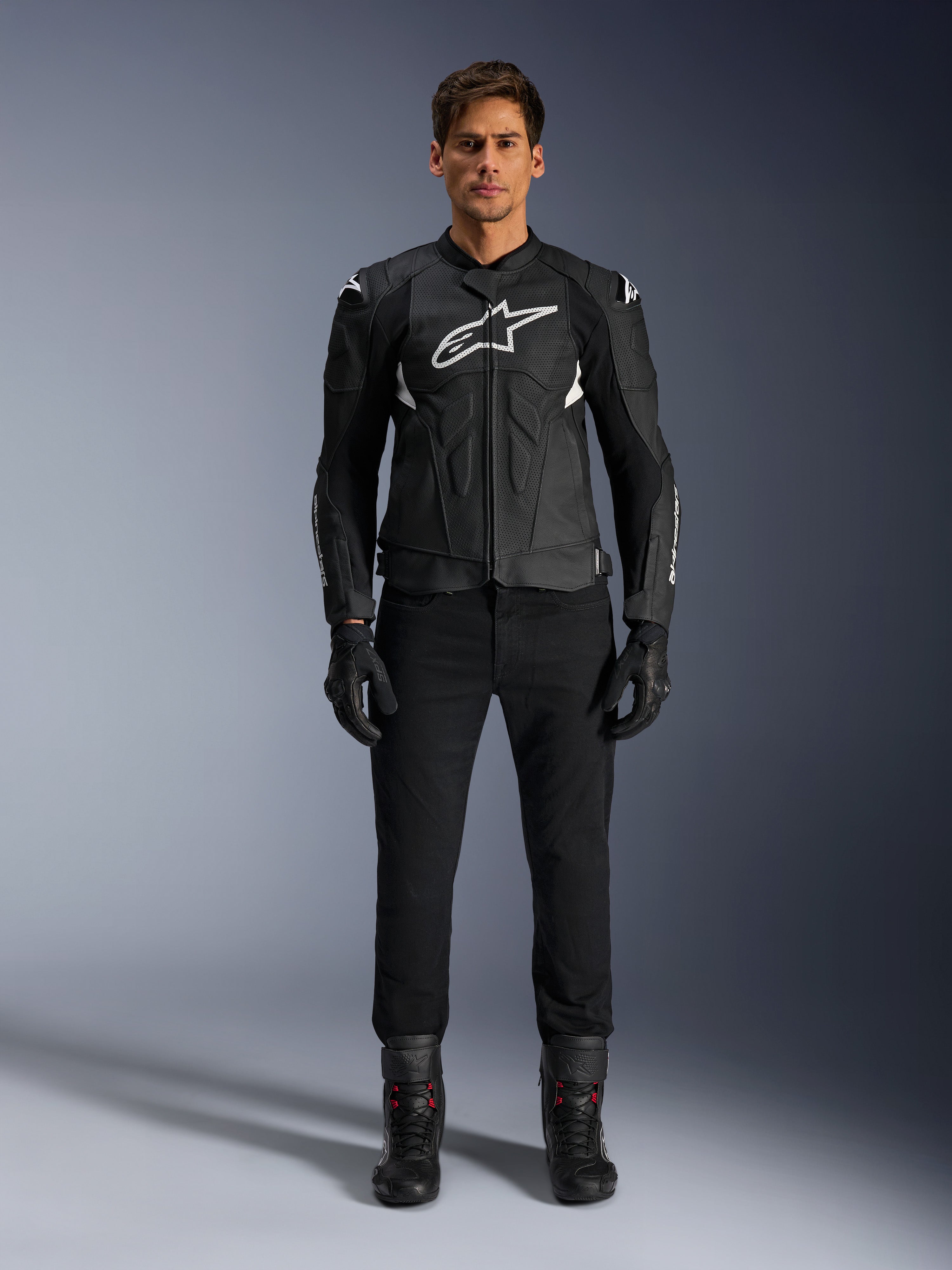 Veste en cuir Alpinestars Dusk Airflow, noir et blanc, veste de moto en cuir avec panneaux perforés pour la circulation de l'air, logo Astars blanc sur la poitrine, portée par un pilote avec pantalon noir, gants et bottes de moto, photo professionnelle en studio pour équipement de moto de sport.