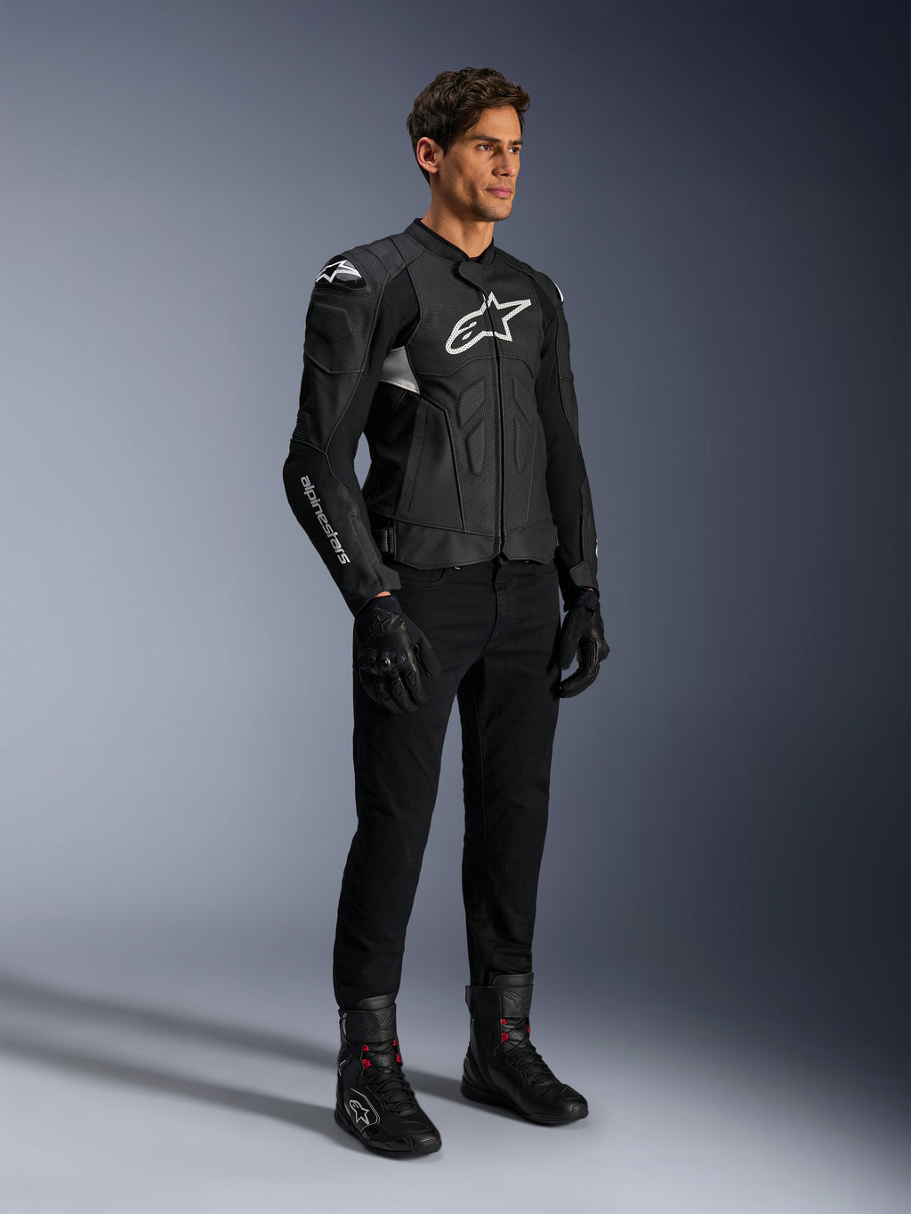 Veste en cuir Alpinestars Dusk Airflow, noir et blanc, portée avec un pantalon de moto, des gants et des bottes noirs, avec panneaux en cuir perforé et logos blancs, style sport road racing