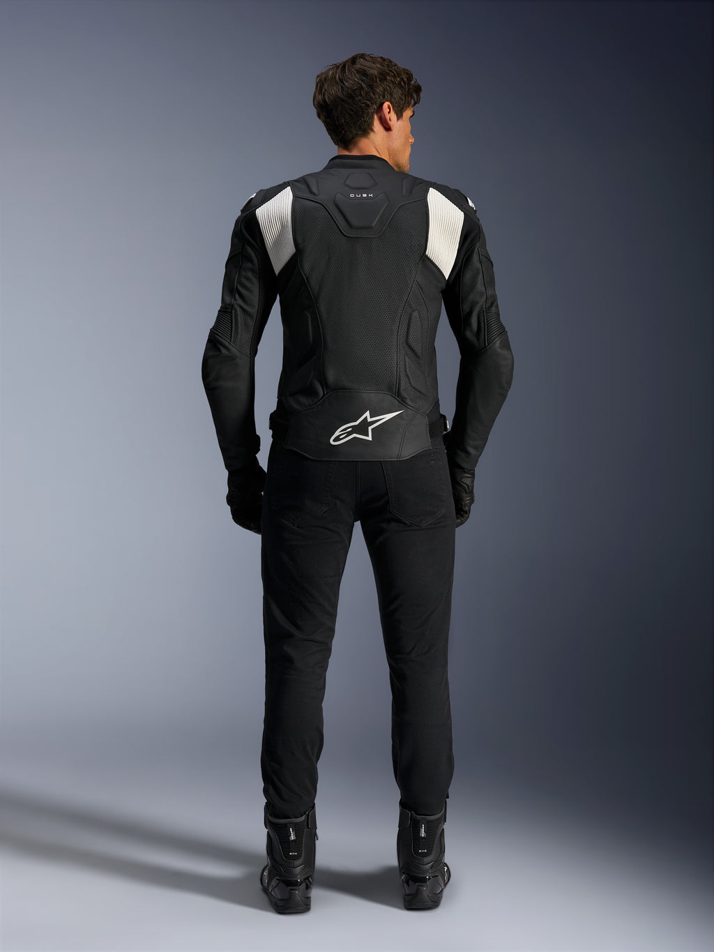 Veste en cuir Alpinestars Dusk Airflow, noir et blanc, vue arrière d'un pilote portant un pantalon et des bottes noirs, dotée de cuir perforé pour la ventilation, de panneaux stretch en accordéon blancs sur les épaules et d'un logo blanc sur le bas du dos