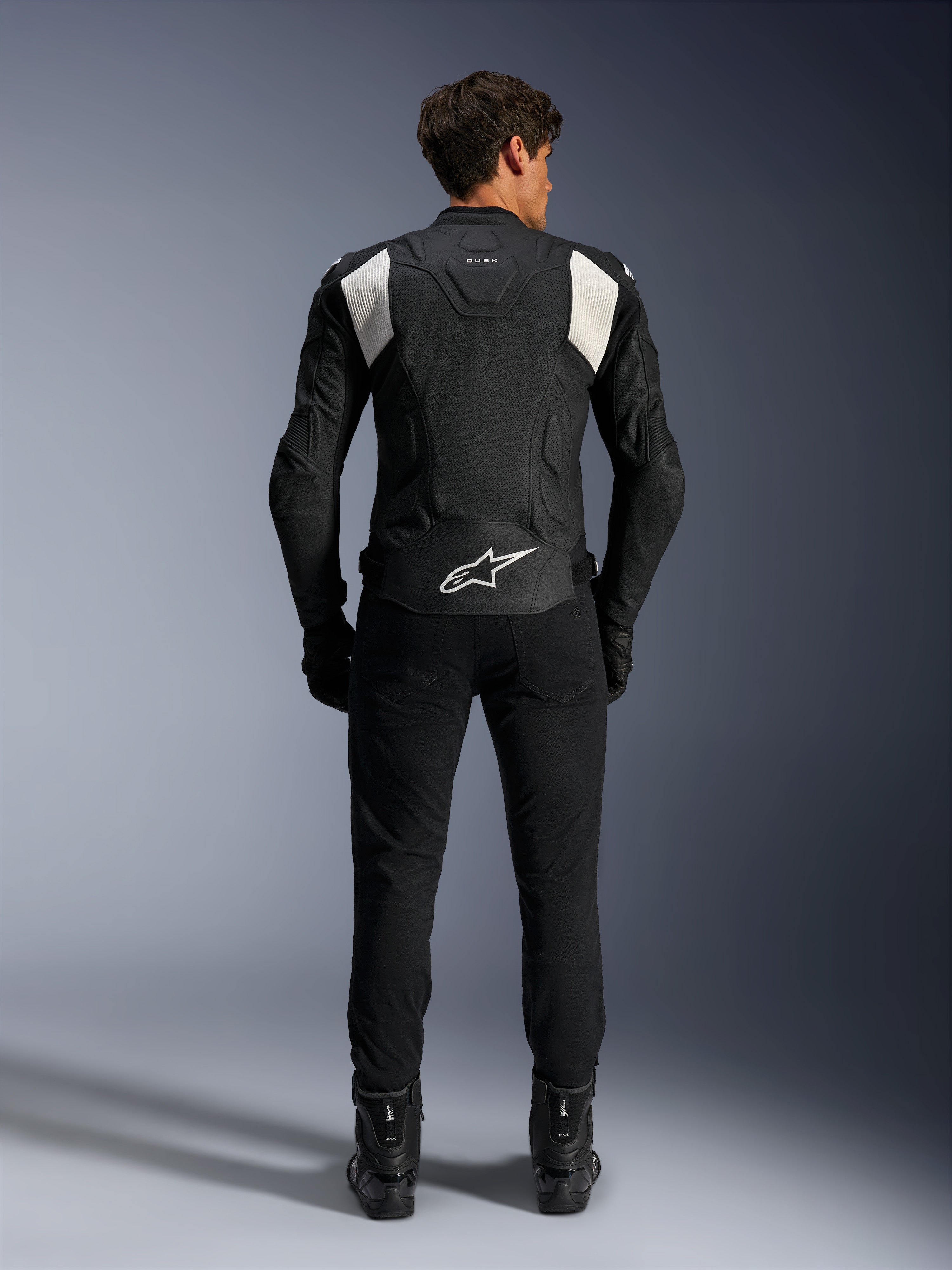 Veste en cuir Alpinestars Dusk Airflow, noir et blanc, vue arrière d'un pilote portant un pantalon et des bottes noirs, dotée de cuir perforé pour la ventilation, de panneaux stretch en accordéon blancs sur les épaules et d'un logo blanc sur le bas du dos