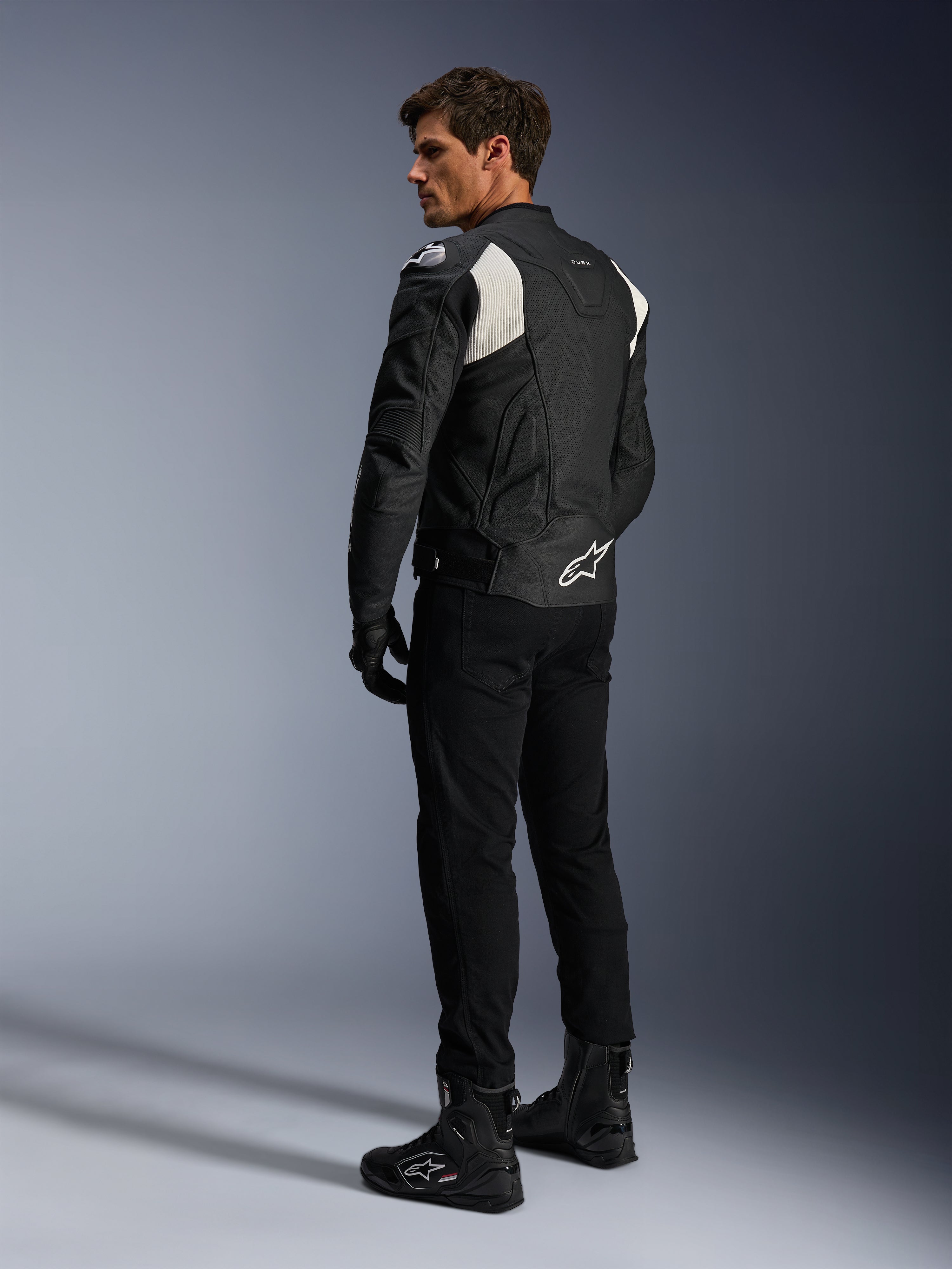 Veste en cuir Alpinestars Dusk Airflow, noir et blanc, vue arrière d'un mannequin portant la veste en cuir perforé avec pantalon et bottes noirs assortis, présentant des panneaux extensibles en accordéon blancs et un logo imposant dans le bas du dos, équipement de moto sport