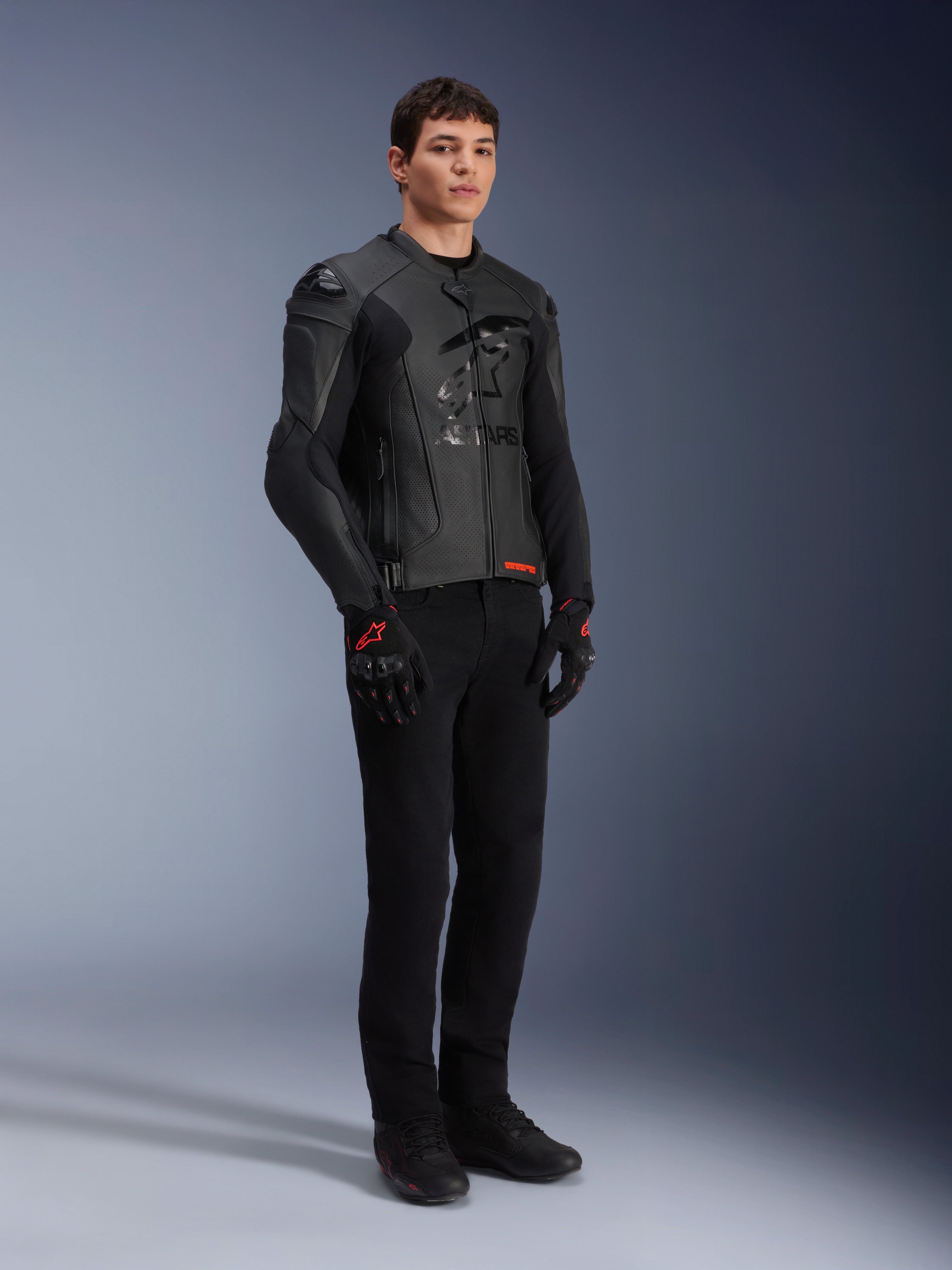 Veste en cuir Alpinestars GP Plus R V4 Airflow, noire, portée par un mannequin homme avec un pantalon de moto noir et des gants aux accents rouges, dotée de panneaux en cuir perforé et de sliders d'épaules intégrés, prise de vue de studio en pied.