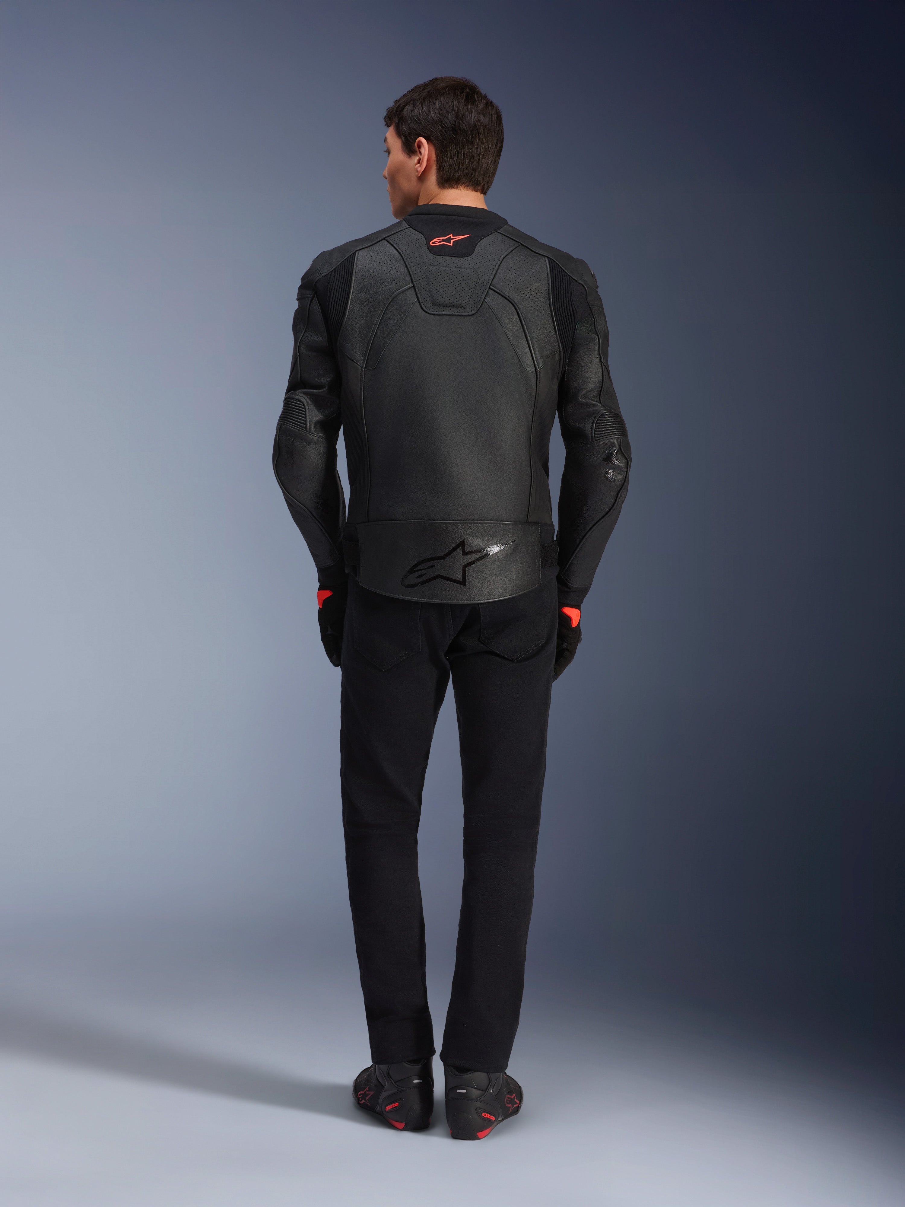 Veste en cuir Alpinestars GP Plus R V4 Airflow, noire, vue arrière du pilote portant l'équipement complet, cuir perforé pour la ventilation et logos rouges, design sportif ajusté, associée à un pantalon noir et des bottes de moto