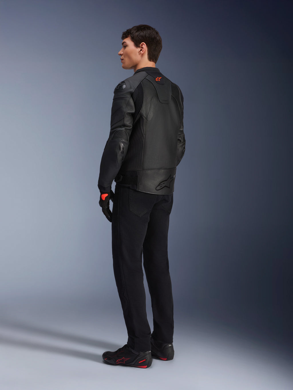 Veste en cuir Alpinestars GP Plus R V4 Airflow, veste de moto sport pour homme, entièrement noire, vue arrière de trois-quarts sur un mannequin, avec panneaux en cuir perforé, logo rouge dans le dos et protections d'épaules intégrées, portée avec un pantalon noir et des chaussures de moto