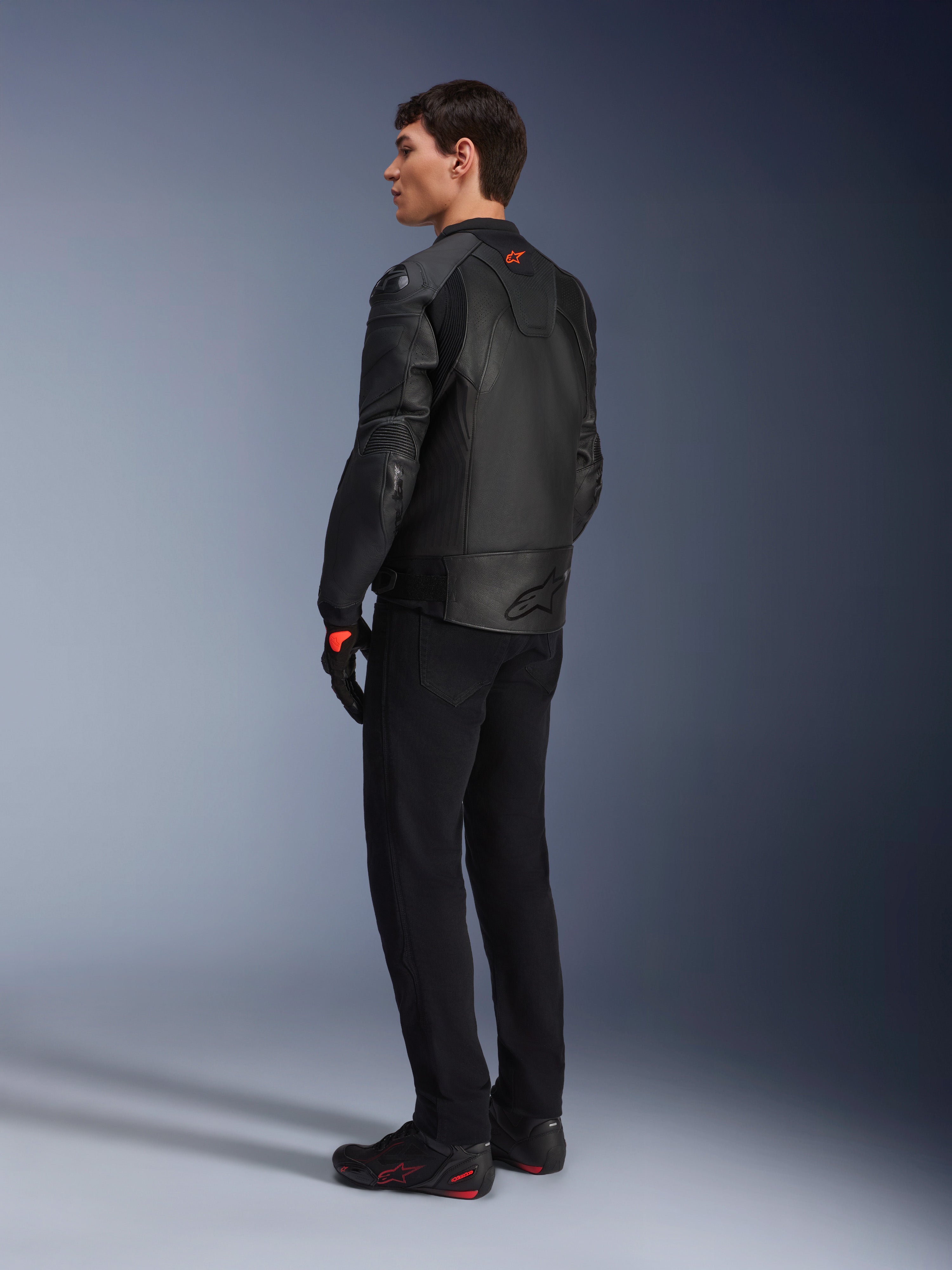 Veste en cuir Alpinestars GP Plus R V4 Airflow, veste de moto sport pour homme, entièrement noire, vue arrière de trois-quarts sur un mannequin, avec panneaux en cuir perforé, logo rouge dans le dos et protections d'épaules intégrées, portée avec un pantalon noir et des chaussures de moto