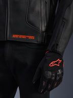 Veste en cuir Alpinestars GP Plus R V4 Airflow, gros plan sur le cuir perforé noir avec texte graphique orange, associée à un gant de moto noir et orange arborant le logo Astars