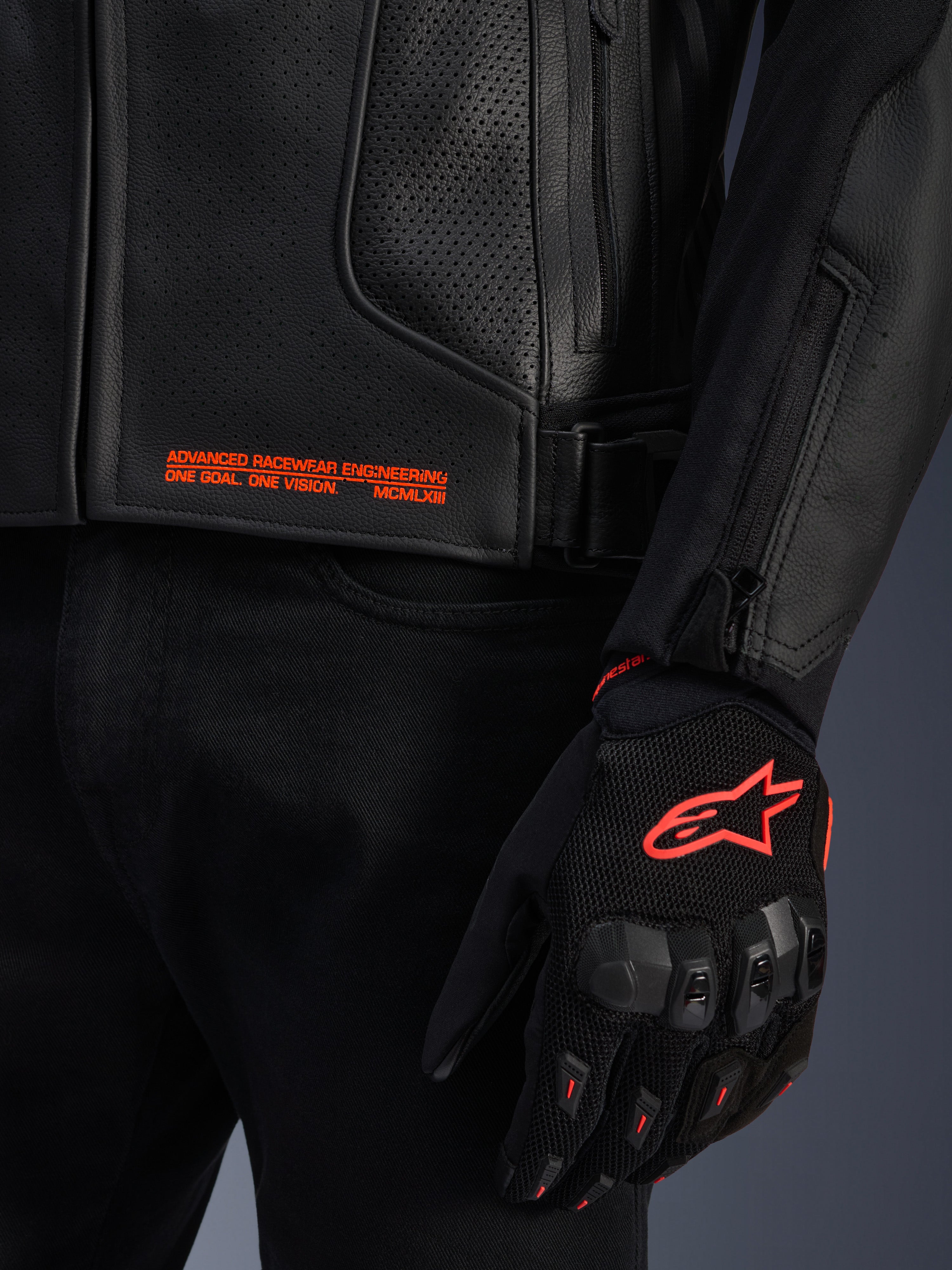 Veste en cuir Alpinestars GP Plus R V4 Airflow, gros plan sur le cuir perforé noir avec texte graphique orange, associée à un gant de moto noir et orange arborant le logo Astars