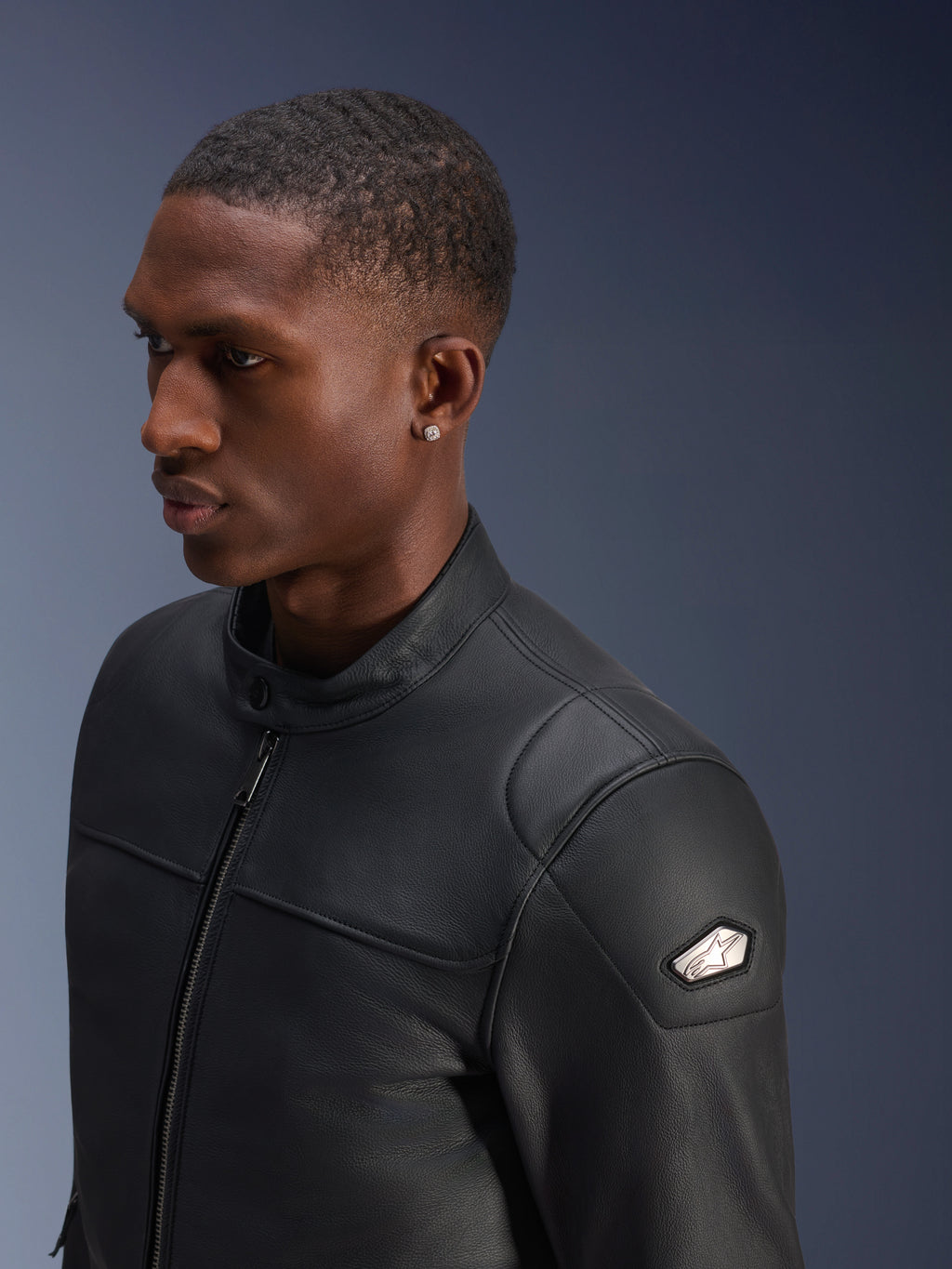 Veste en cuir Alpinestars Blacktrack, cuir de bovin noir premium, vue de profil en gros plan d'un homme portant la veste, fermeture éclair frontale métallique canon de fusil, col haut boutonné, écusson d'épaule métallique avec logo, veste de moto élégante à la coupe ajustée
