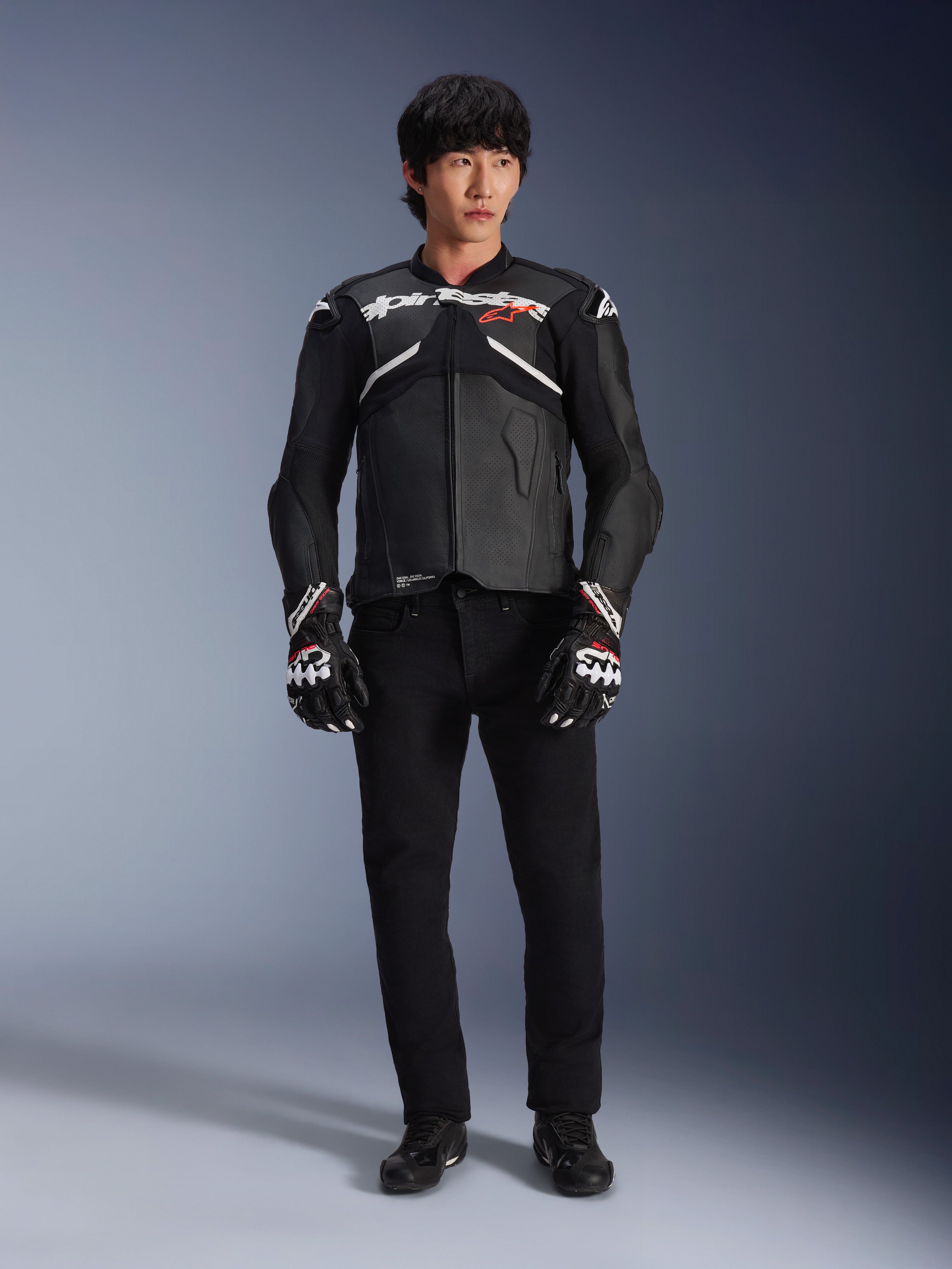 Mannequin portant la veste en cuir Alpinestars Atem V5 en noir et blanc, avec le logo Alpinestars blanc et un logo d'étoile rouge sur la poitrine, associée à un pantalon noir et des gants de course noir et blanc, équipement de moto performant et élégant