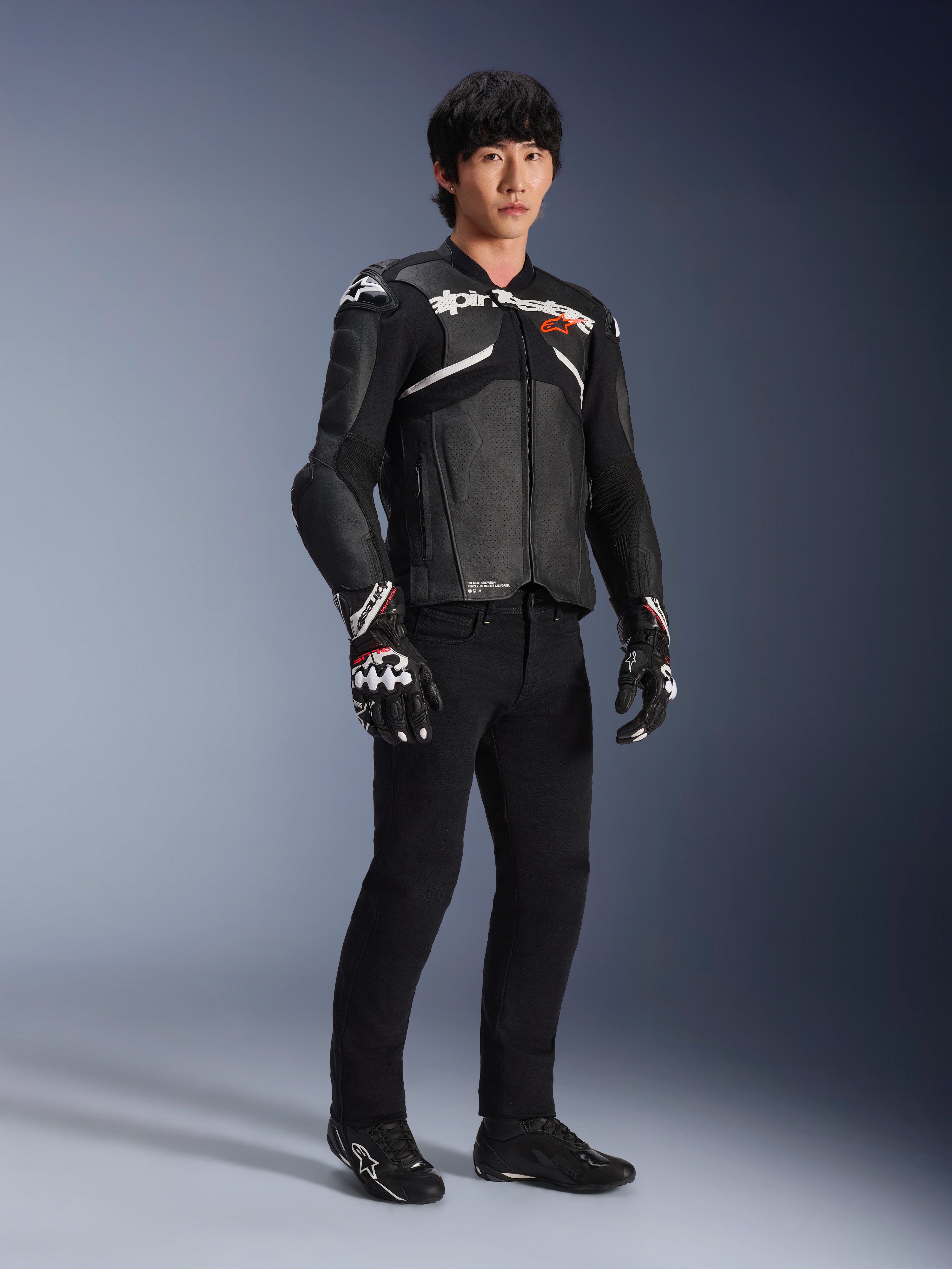 Veste en cuir Atem V5 Alpinestars, veste de course moto, noir et blanc, portée avec un pantalon noir et des gants assortis, avec le logo Alpinestars blanc sur la poitrine et des protections aux épaules.