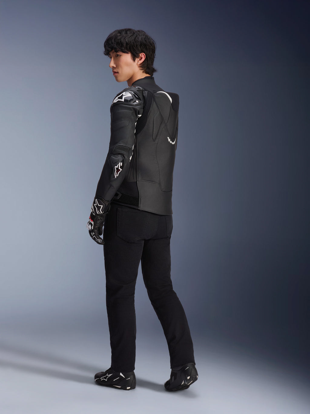 Modèle masculin portant la veste en cuir Alpinestars Atem V5 en noir et blanc, vue de trois-quarts arrière montrant la bosse aérodynamique et les protections DFS RR, assortie d'un pantalon noir, de gants de course et de bottes, équipement de moto de course professionnel
