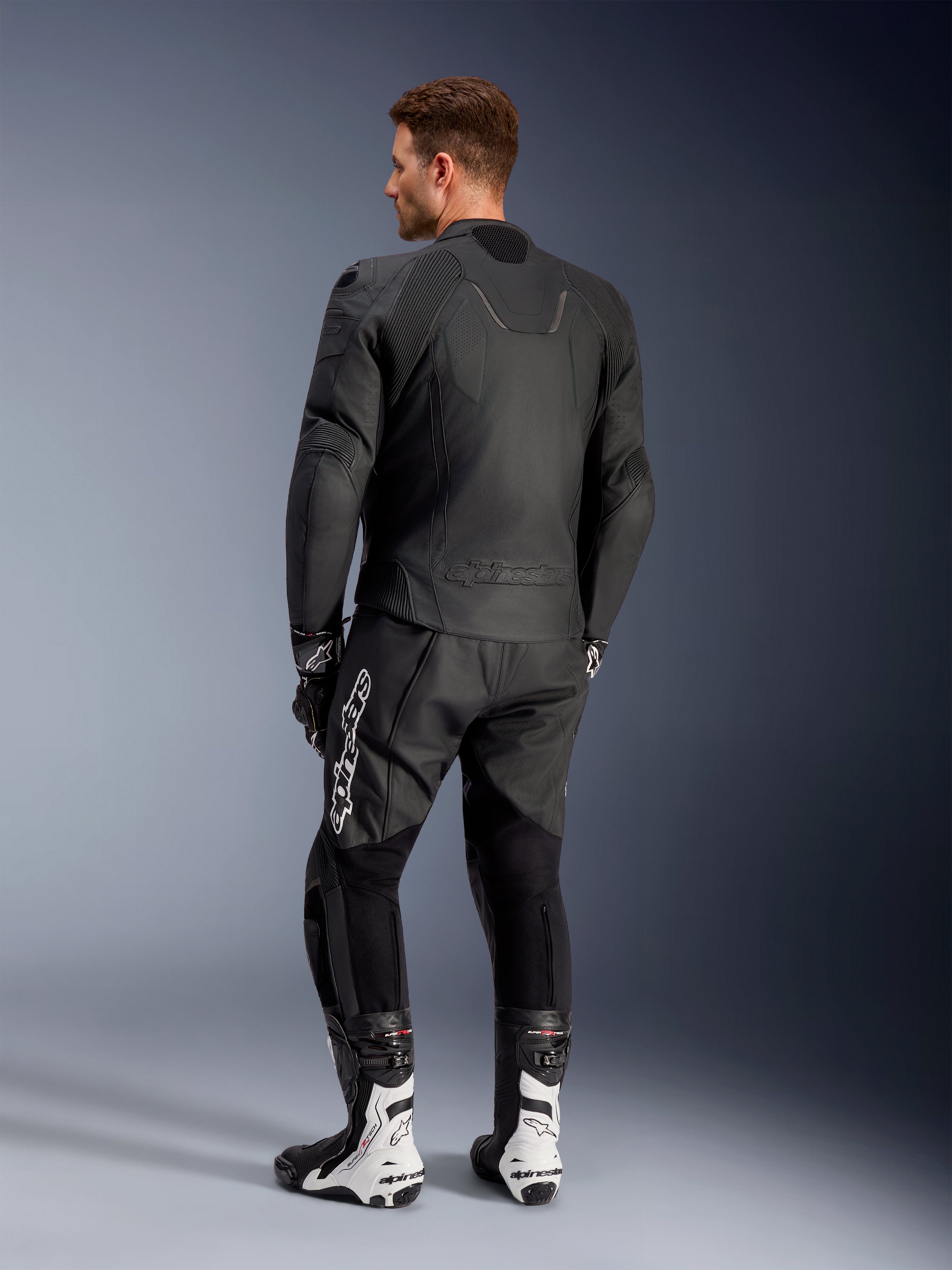 Veste en cuir Alpinestars Caliber, blouson de moto de route noir, vue de dos, avec cuir perforé et inserts stretch accordéon à la taille et aux épaules, logo en relief, porté avec un pantalon en cuir noir et des bottes de course blanches