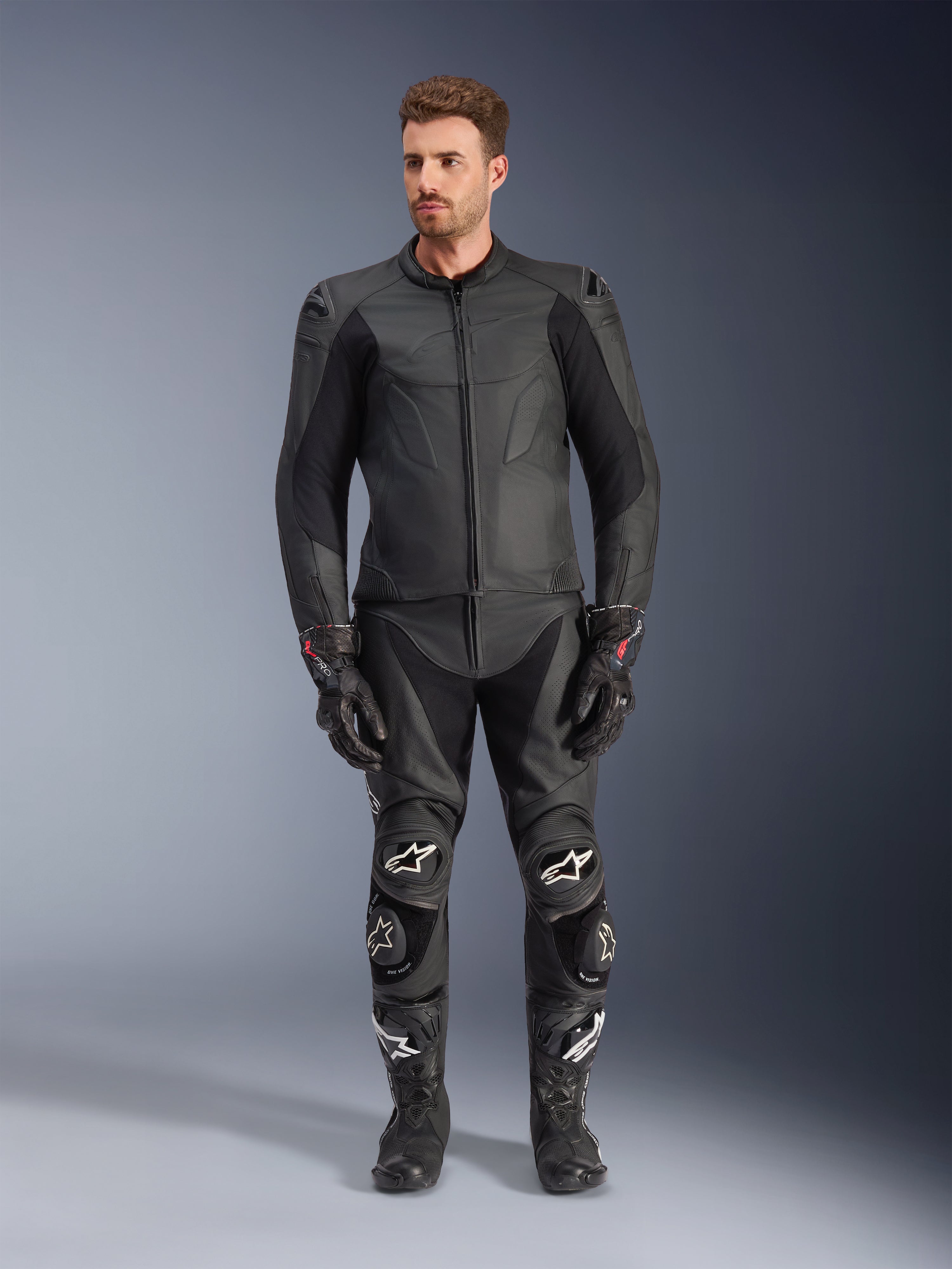 Veste en cuir Alpinestars Caliber, veste de moto noire, portée avec un pantalon de course, des gants et des bottes assortis, dotée d'un design épuré avec des logos ton sur ton, des panneaux stretch sur les manches et des sliders de protection aux épaules, cadre de studio professionnel