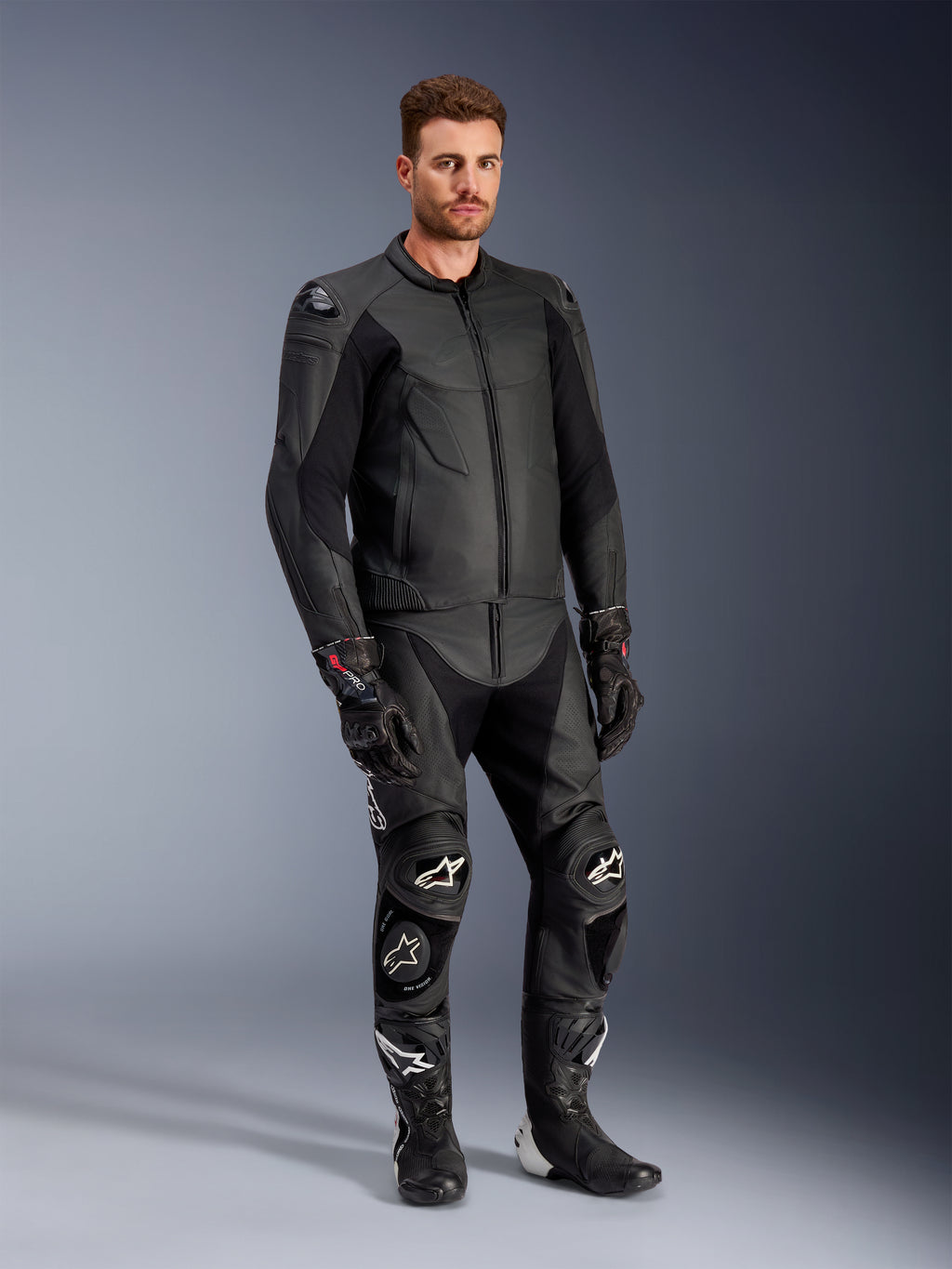 Modèle masculin portant la veste en cuir Alpinestars Caliber noire, associée à un pantalon de moto en cuir noir assorti, des gants de course et des bottes, présentant une tenue complète d'équipement de route sportif.