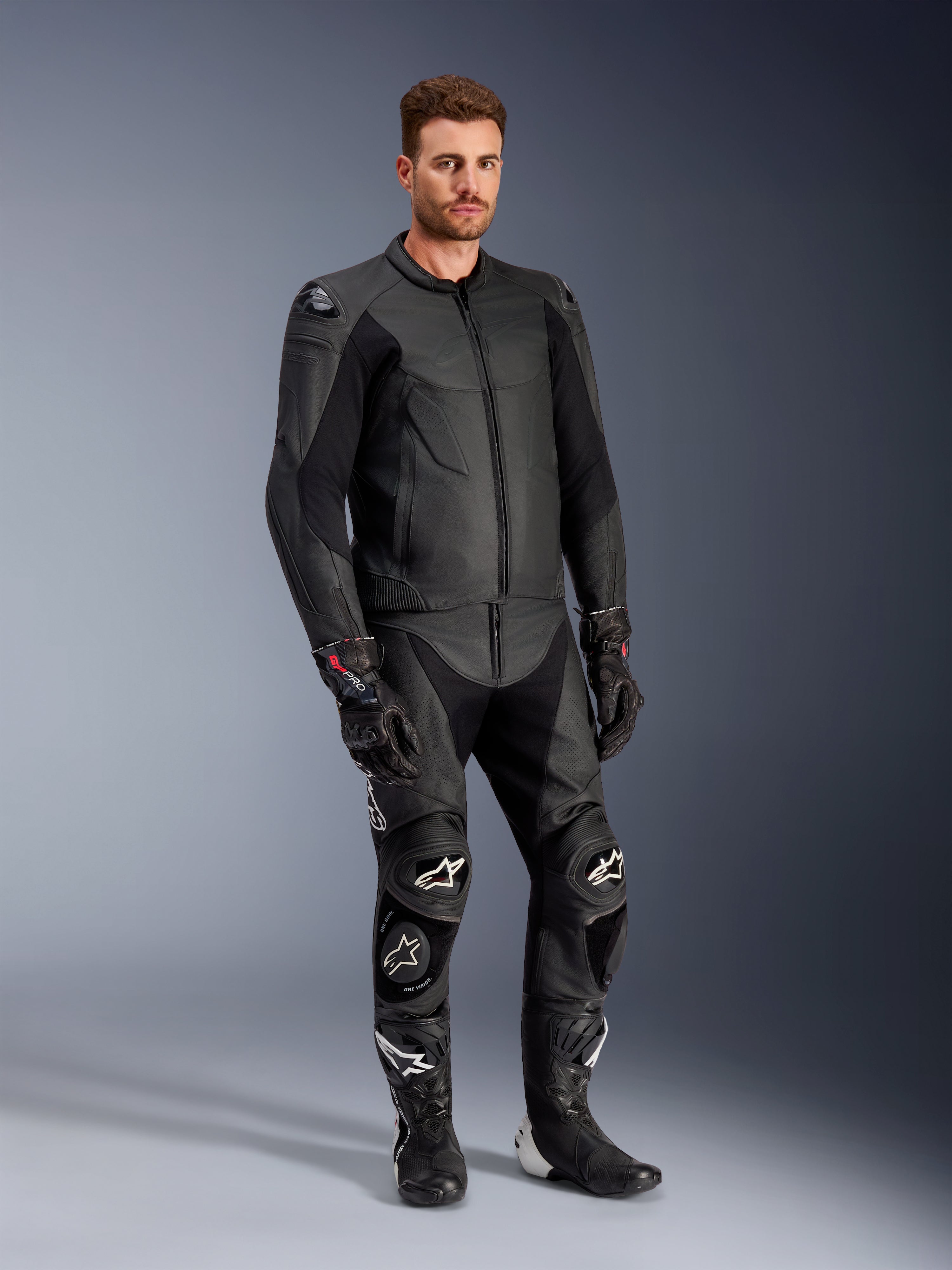 Modèle masculin portant la veste en cuir Alpinestars Caliber noire, associée à un pantalon de moto en cuir noir assorti, des gants de course et des bottes, présentant une tenue complète d'équipement de route sportif.