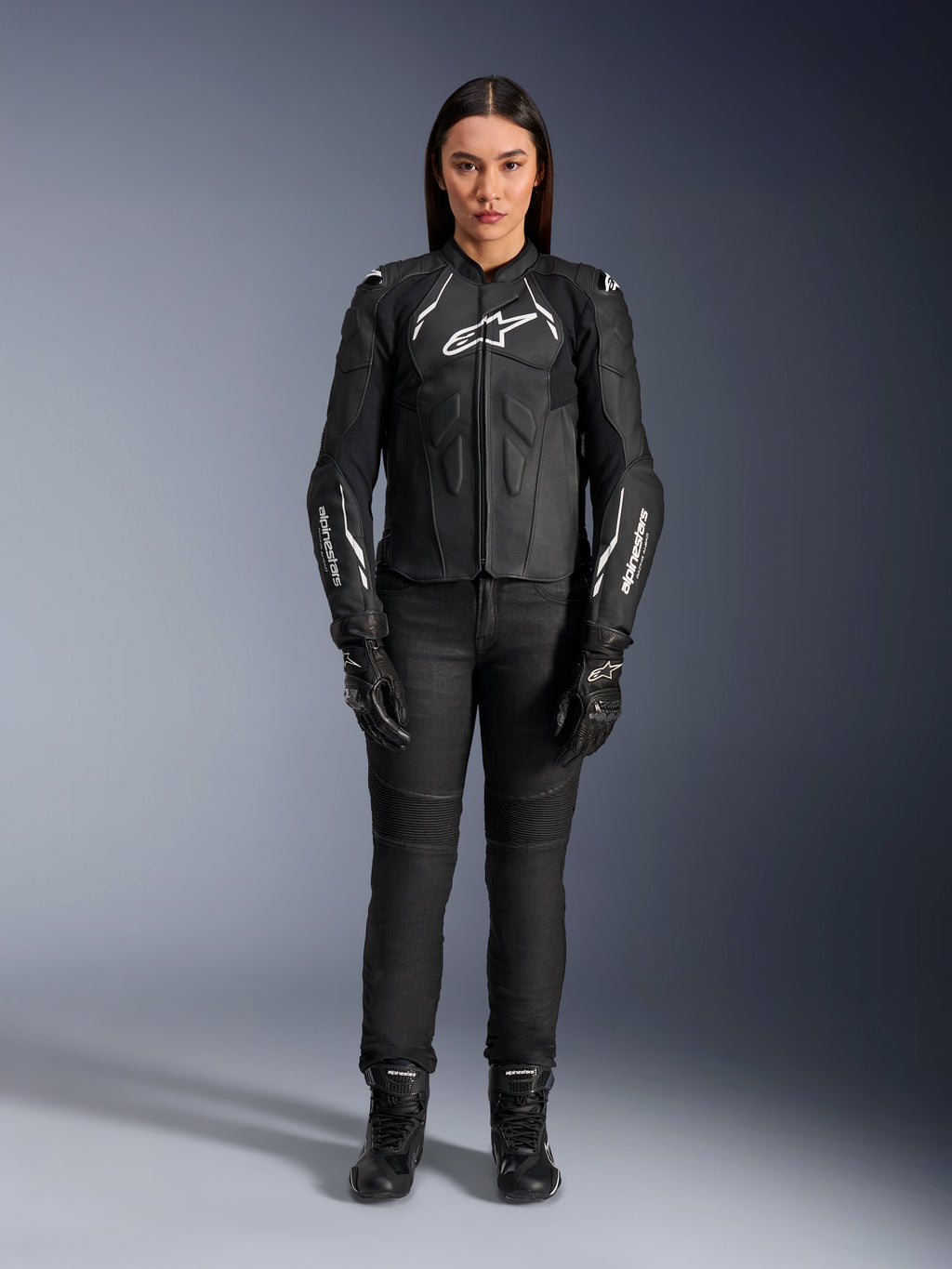 Veste en cuir pour femme Alpinestars Stella Dusk, noir et blanc, portée par un mannequin avec un pantalon et des bottes noirs assortis, vue de face, logos Alpinestars blancs sur la poitrine et les manches, équipement de protection en cuir bovin premium pour femme