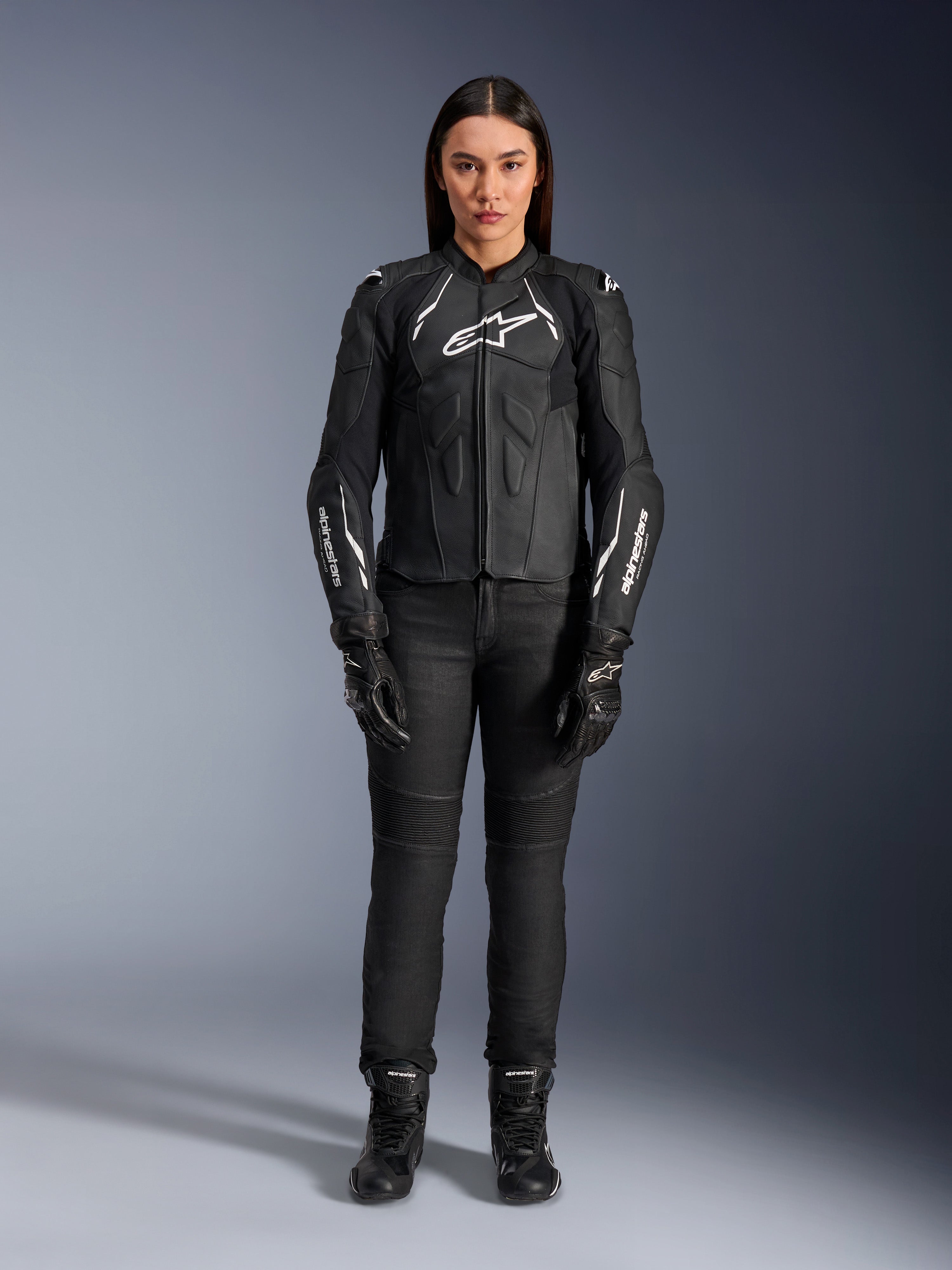 Veste en cuir pour femme Alpinestars Stella Dusk, noir et blanc, portée par un mannequin avec un pantalon et des bottes noirs assortis, vue de face, logos Alpinestars blancs sur la poitrine et les manches, équipement de protection en cuir bovin premium pour femme