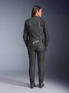 Veste en cuir pour femme Stella Dusk d'Alpinestars, noire, vue arrière avec logo Alpinestars blanc en bas du dos et marquage 'DUSK' en haut du dos, portée avec un pantalon de moto et des bottes noirs, mettant en valeur la coupe féminine ajustée et la construction protectrice en cuir.