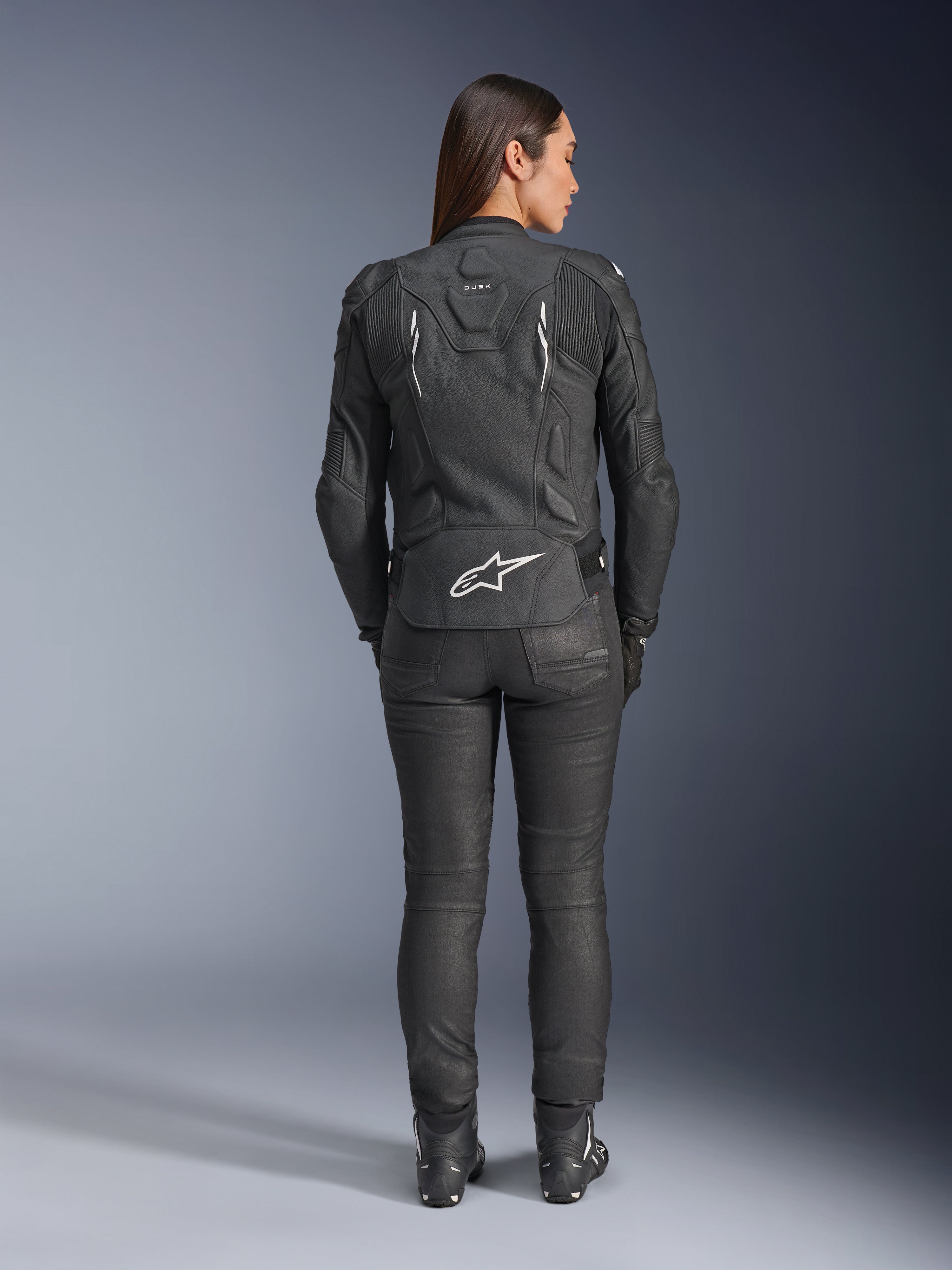 Veste en cuir pour femme Stella Dusk d'Alpinestars, noire, vue arrière avec logo Alpinestars blanc en bas du dos et marquage 'DUSK' en haut du dos, portée avec un pantalon de moto et des bottes noirs, mettant en valeur la coupe féminine ajustée et la construction protectrice en cuir.