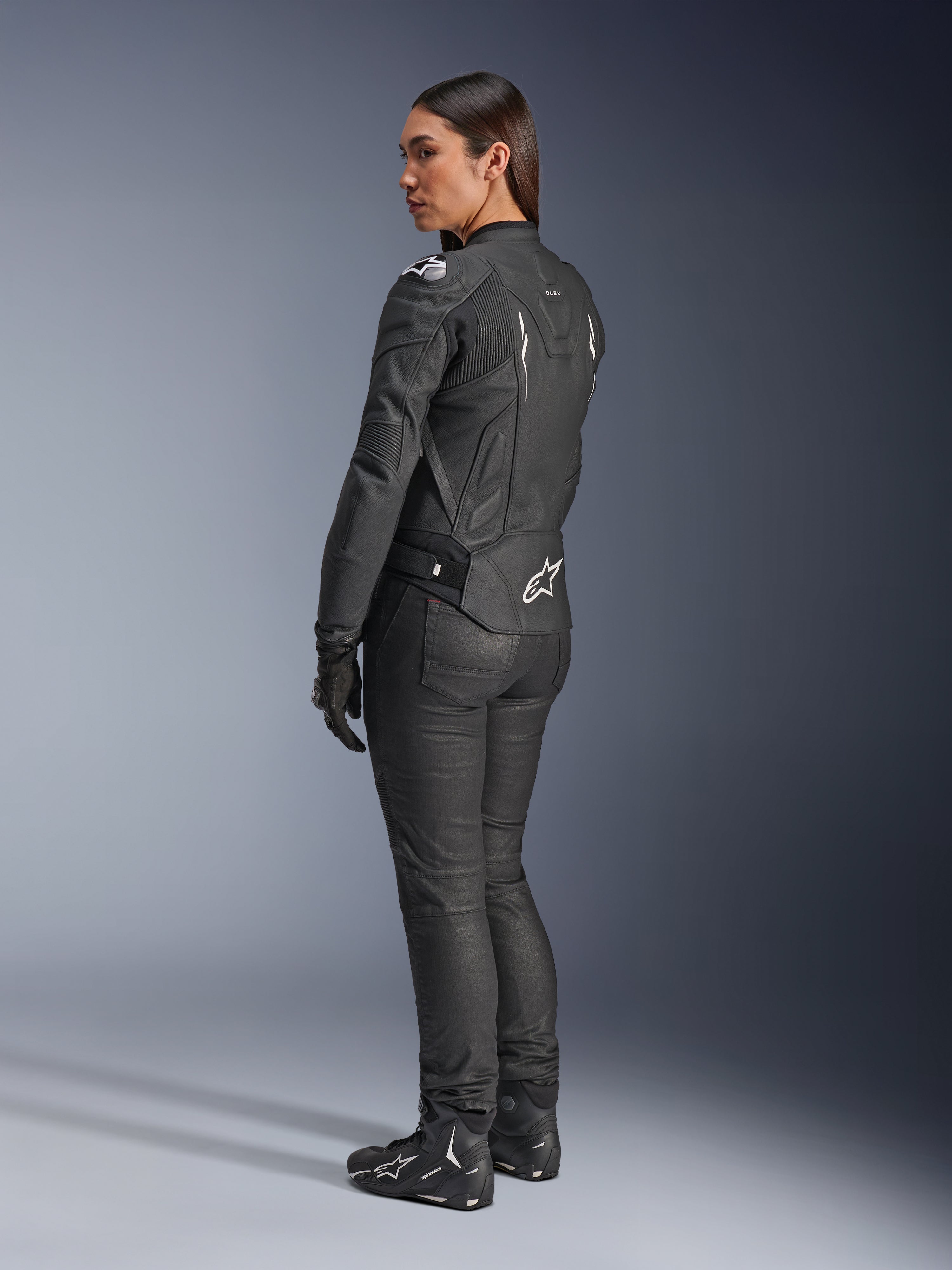 Veste en cuir Alpinestars Stella Dusk, veste de moto pour femme, noir et blanc, vue arrière présentant le logo Alpinestars blanc sur le bas du dos et les épaules, portée avec un pantalon de moto noir et des bottes, coupe ergonomique axée sport pour la conduite sur route