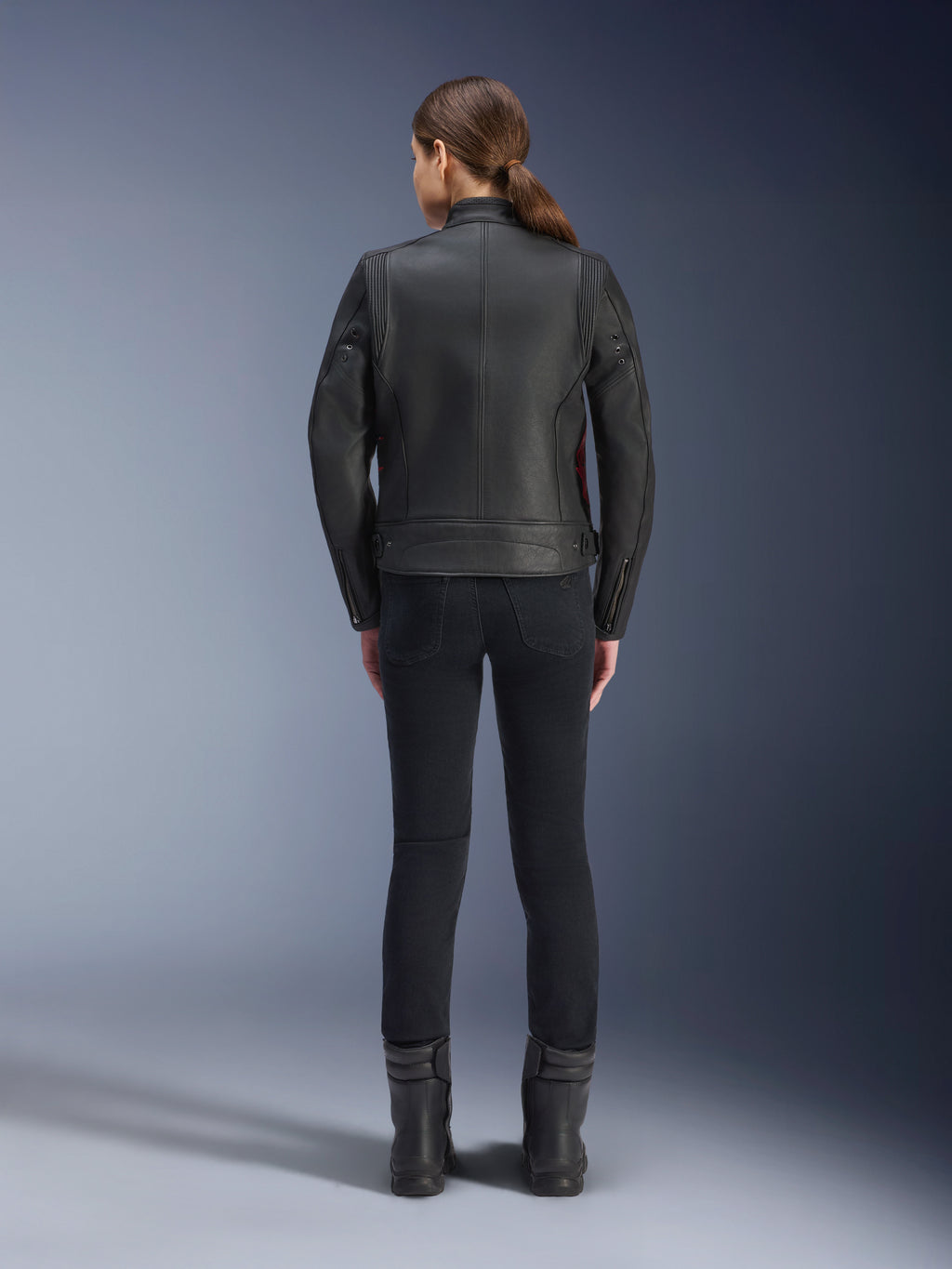 Vue arrière d'une femme portant la veste en cuir Alpinestars Stella Dyno, noir avec accents rouge Haute, associée à un pantalon noir ajusté et des bottes noires, design de veste de moto héritage moderne.
