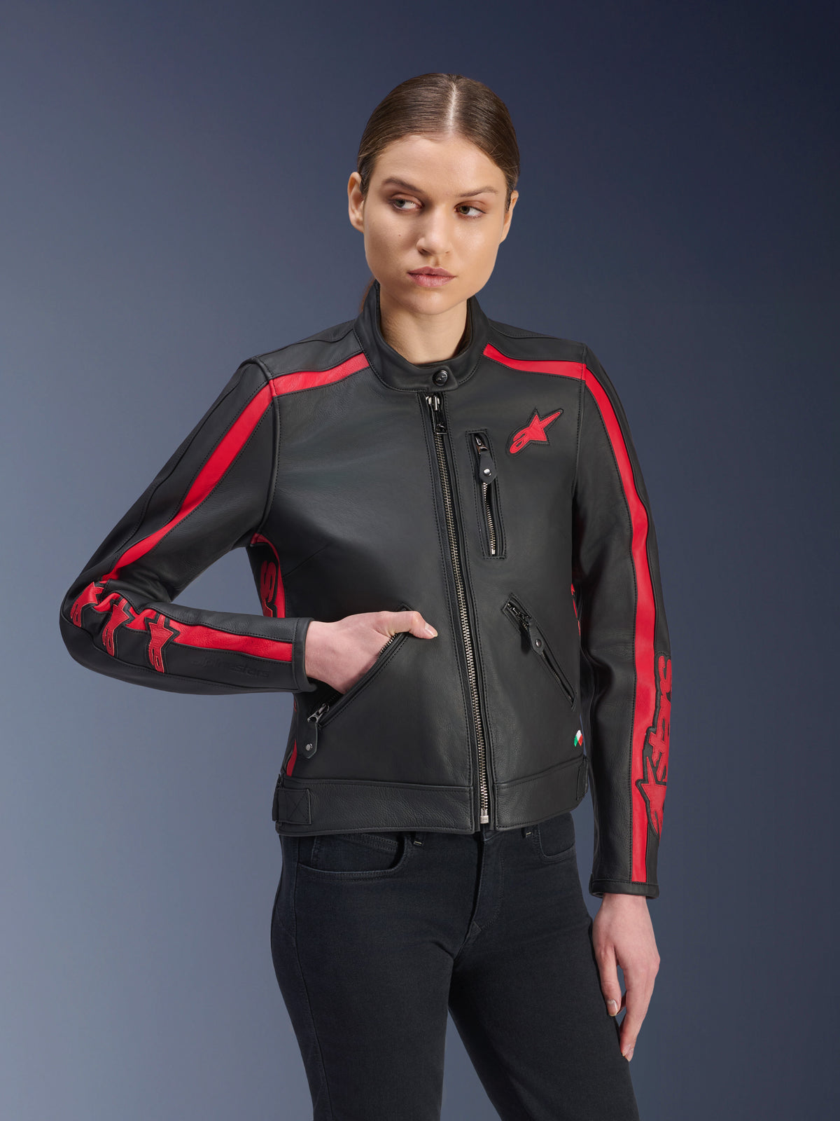 Veste en cuir Stella Dyno Alpinestars, veste de moto pour femme, noir et rouge Haute, portée par un mannequin avec un jean noir, bandes de course rouges sur les épaules et les manches avec logo distinctif, conception en cuir haut de gamme, design d'inspiration vintage pour la conduite urbaine