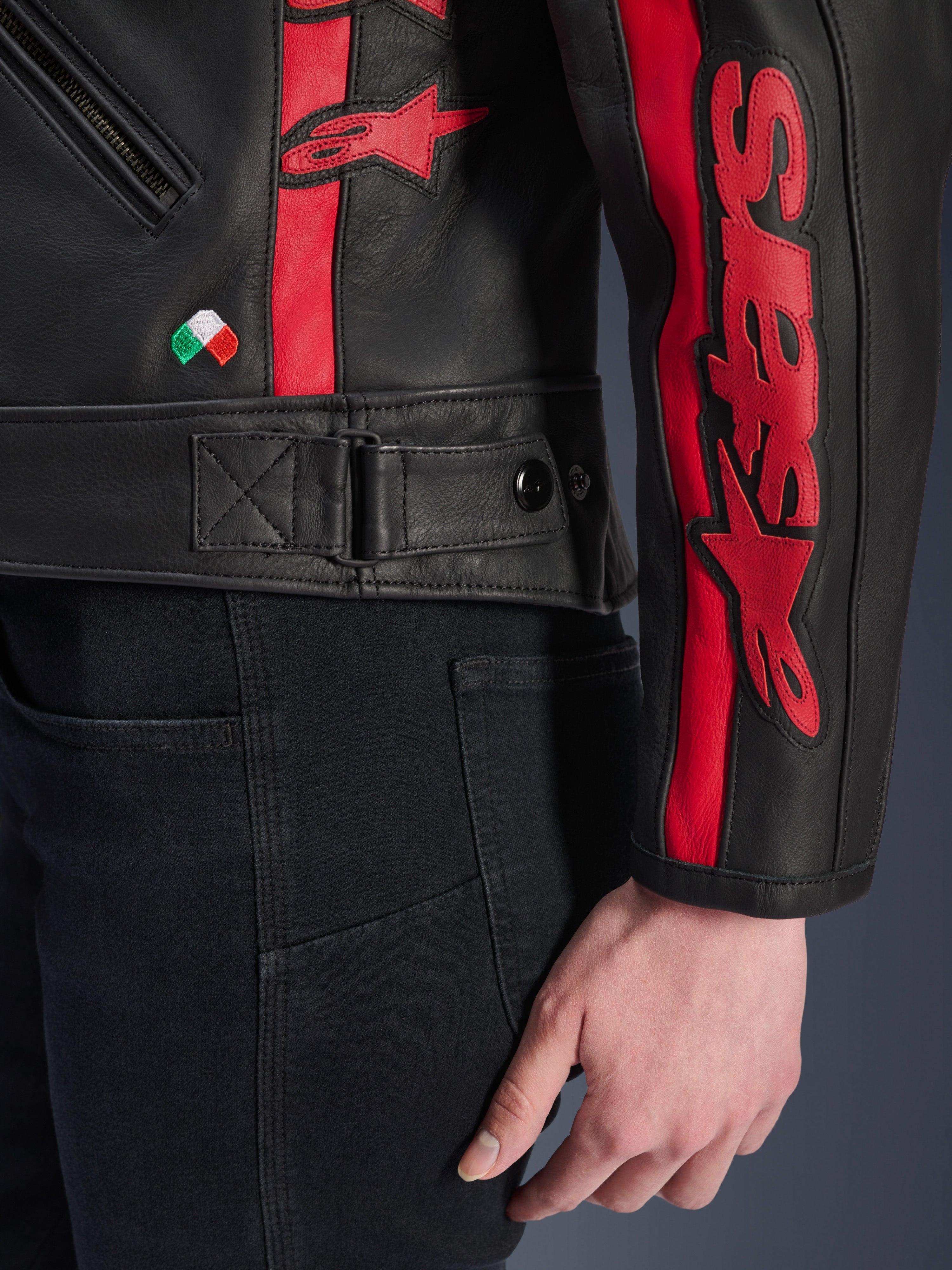 Veste en cuir Stella Dyno d'Alpinestars, veste de moto en cuir pour femme, noir et rouge Haute Red, vue rapprochée de la taille et de la manche, bandes de course rouges avec logo Alpinestars, sangle de réglage à la taille, broderie du petit drapeau italien, construction en cuir de bovin de qualité supérieure