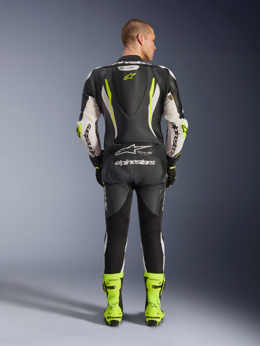 Combinaison en cuir Alpinestars GP Tech V5 1PC, vue de dos, noir, blanc et jaune fluo, avec bosse aérodynamique et logo étoile jaune, marquage Alpinestars blanc sur les manches et le dos, bottes de course jaune fluo assorties, équipement de moto professionnel pour circuit