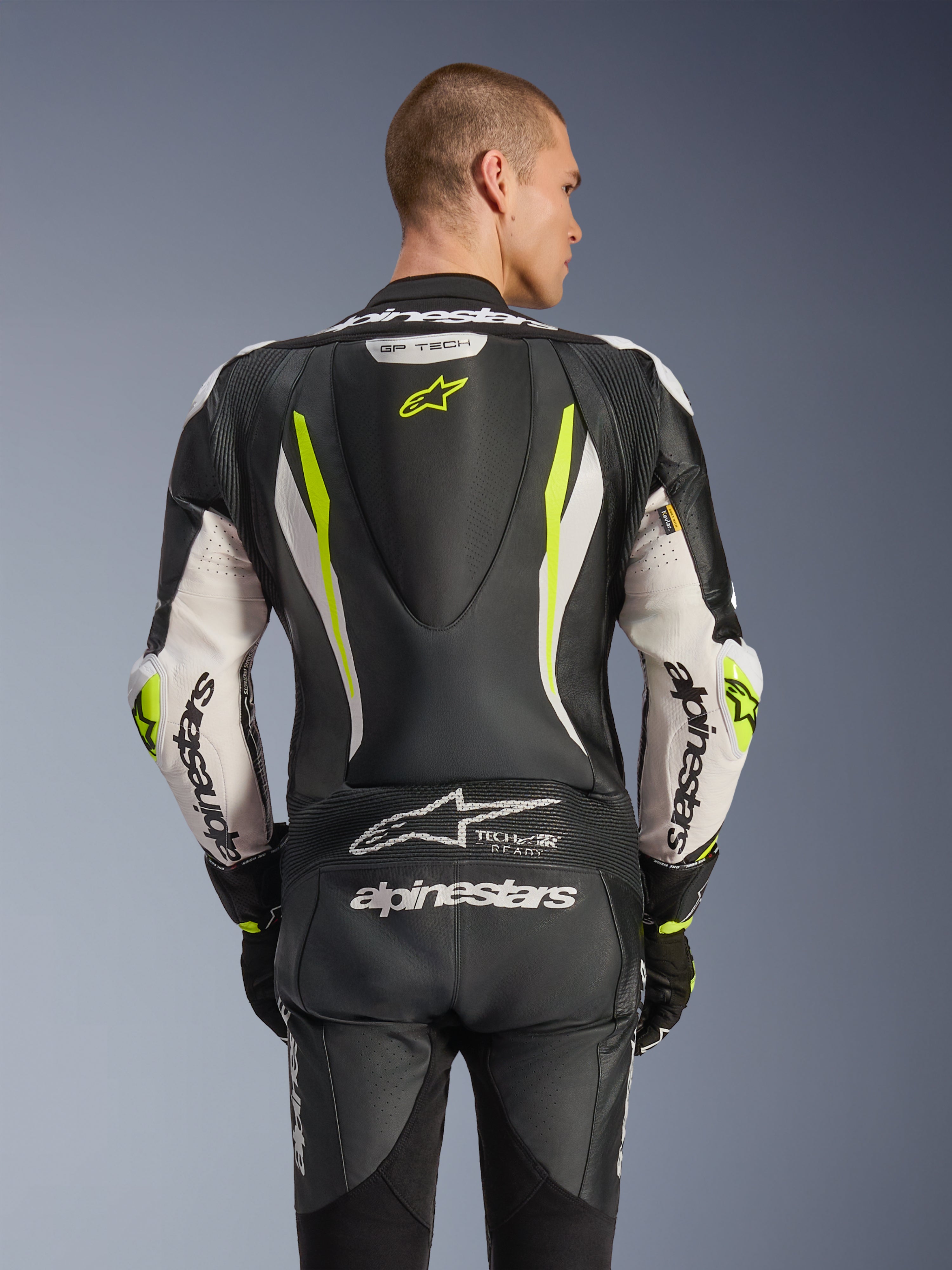 Combinaison en cuir Alpinestars GP Tech V5 une pièce, vue de dos sur mannequin, Noir, Blanc et Jaune Fluo, dotée d'une bosse aérodynamique, d'un large marquage Alpinestars sur les bras et l'assise, de panneaux extensibles en accordéon et du logo Tech-Air Ready