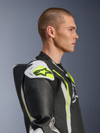 Modèle masculin portant la combinaison en cuir Alpinestars GP Tech V5 1PC en Noir Blanc Jaune Fluo, vue de profil mettant en valeur la bosse de vitesse aérodynamique et les sliders d'épaule blancs, équipement de protection moto inspiré de la course avec des détails en cuir perforé