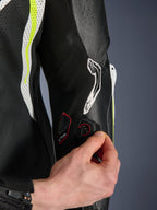 Combinaison en cuir GP Tech V5 une pièce, vue détaillée du port de connexion Tech-Air sur la manche, main démontrant l'intégration électronique, cuir noir avec panneaux perforés jaune fluo et blanc, équipement de course professionnel