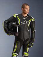 Combinaison en cuir Alpinestars GP Tech V5 une pièce, noir, blanc et jaune fluo, mannequin masculin tenant un casque de moto noir, avec panneaux perforés, accents blancs et jaune fluo, marquage Alpinestars proéminent, équipement de course moto professionnel