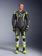 Alpinestars GP Tech V5 One Piece Leather Suit, combinaison de course moto professionnelle en noir et jaune fluo avec accents blancs, portée par un mannequin avec des gants et des bottes de course assortis, dotée d'un design aérodynamique et de sliders de protection.
