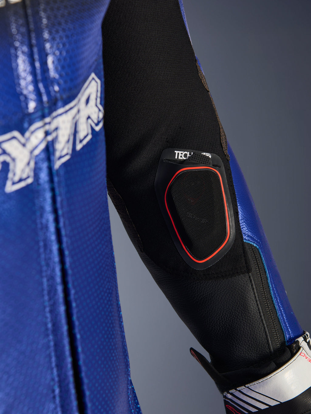 Close-up detail of the Yamaha GP-R7 One Piece Leather Suit forearm, featuring the integrated Tech-Air 10 Bluetooth display pocket with red trim, metallic blue perforated leather, and black stretch fabric inserts for racing performance.