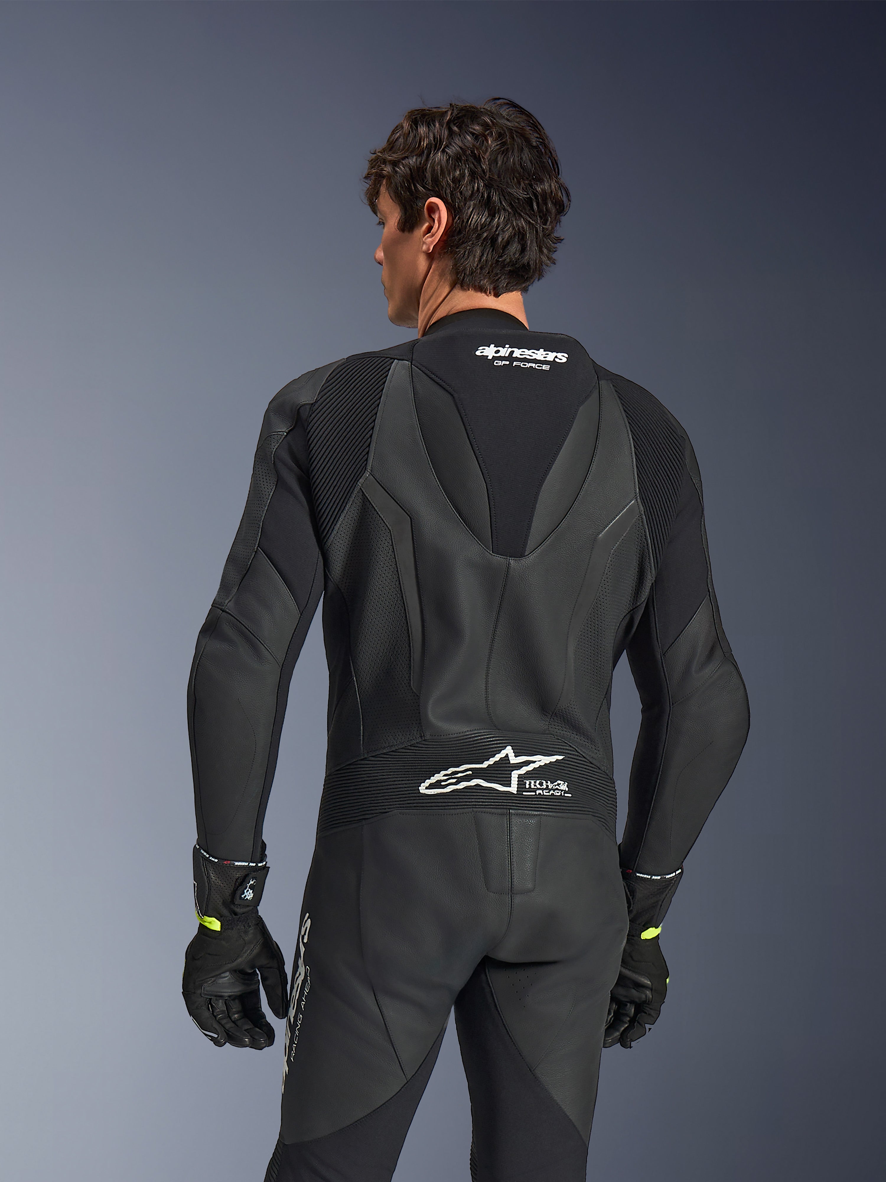 Combinaison en cuir Alpinestars GP Force V2, une pièce, noire, vue de dos montrant le cuir perforé et les panneaux extensibles en accordéon, logos Alpinestars et Tech-Air Ready blancs sur le haut et le bas du dos, portée avec des gants noirs, équipement de course moto haute performance