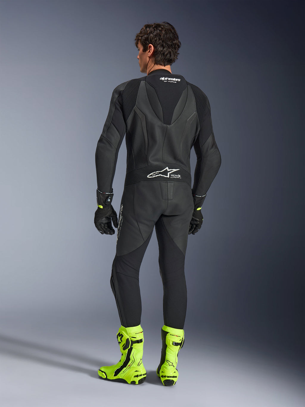 Combinaison en cuir Alpinestars GP Force V2 - 1 pièce, noir, vue de dos portée par un mannequin, avec bosse aérodynamique et marquage Tech-Air Ready, assortie de gants noirs et de bottes de course jaune fluorescent vif, équipement de piste professionnel