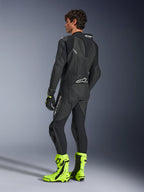 Combinaison en cuir Alpinestars GP Force V2, combinaison de course moto 1 pièce noire, vue arrière montrant la bosse aérodynamique et le cuir perforé, portée avec des bottes de course jaune fluo et des gants noirs, équipement professionnel prêt pour la piste