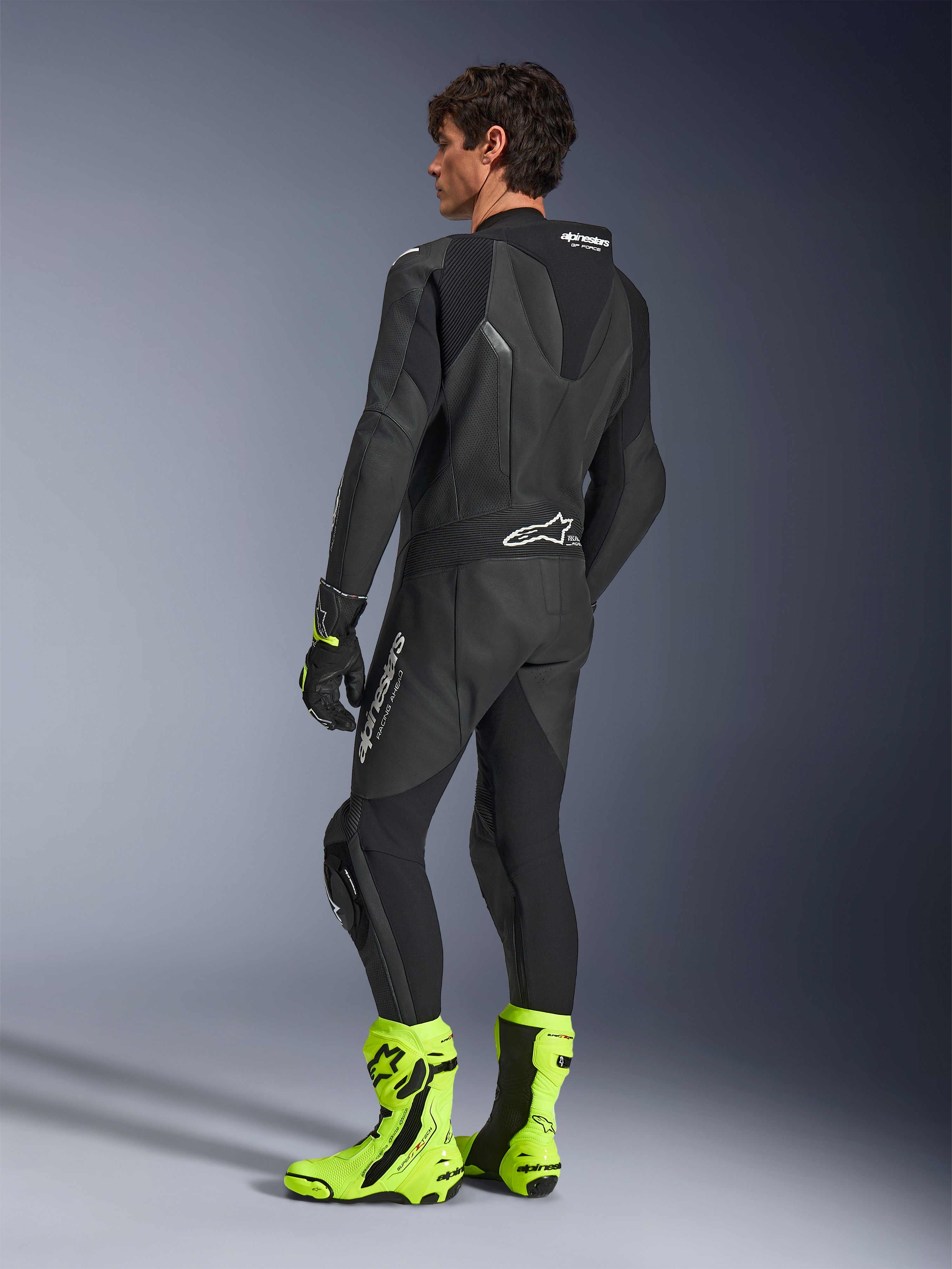 Combinaison en cuir Alpinestars GP Force V2, combinaison de course moto 1 pièce noire, vue arrière montrant la bosse aérodynamique et le cuir perforé, portée avec des bottes de course jaune fluo et des gants noirs, équipement professionnel prêt pour la piste