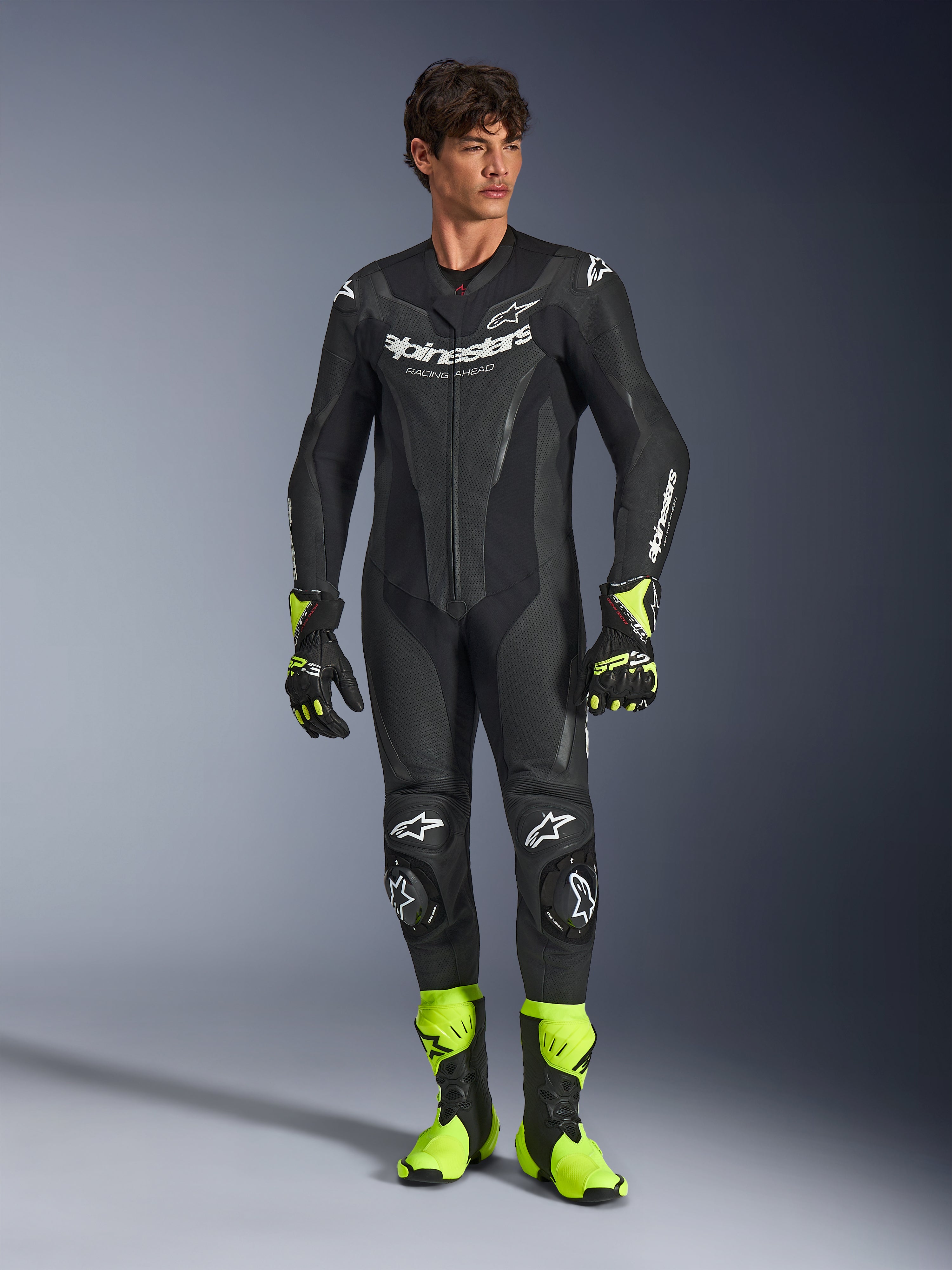 Combinaison en cuir Alpinestars GP Force V2, combinaison de course moto une pièce noire, portée avec des bottes et des gants jaune fluo, dotée de logos blancs et de cuir perforé, design ajusté pour les journées sur piste