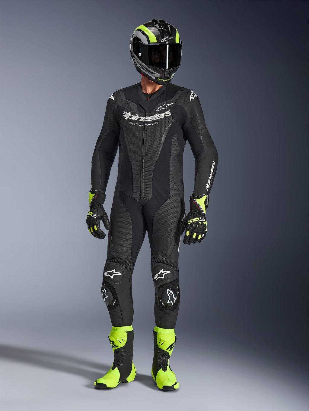 Pilote de moto portant la combinaison en cuir Alpinestars GP Force V2 noire, combinaison une pièce avec logos blancs, accompagnée d'un casque, de gants et de bottes de course noir et jaune fluo, équipement de piste professionnel sur fond de studio gris
