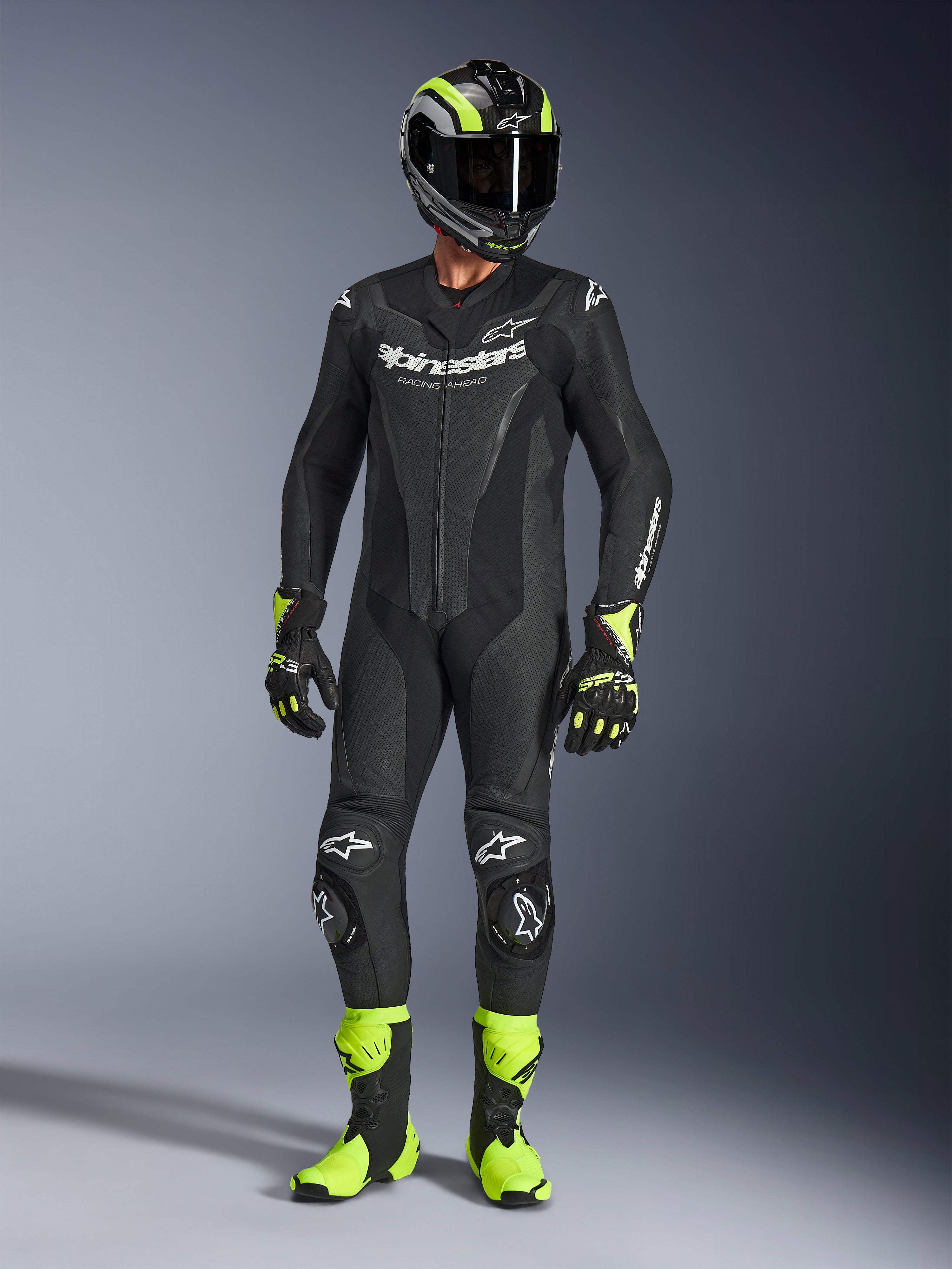 Pilote de moto portant la combinaison en cuir Alpinestars GP Force V2 noire, combinaison une pièce avec logos blancs, accompagnée d'un casque, de gants et de bottes de course noir et jaune fluo, équipement de piste professionnel sur fond de studio gris