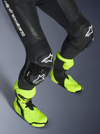 Combinaison en cuir Alpinestars GP Force V2, combinaison une pièce en cuir noir, gros plan sur les jambes et les genoux avec sliders à logo blanc, panneaux extensibles en accordéon, portée avec des bottes de course jaune fluo, équipement de course moto haute performance
