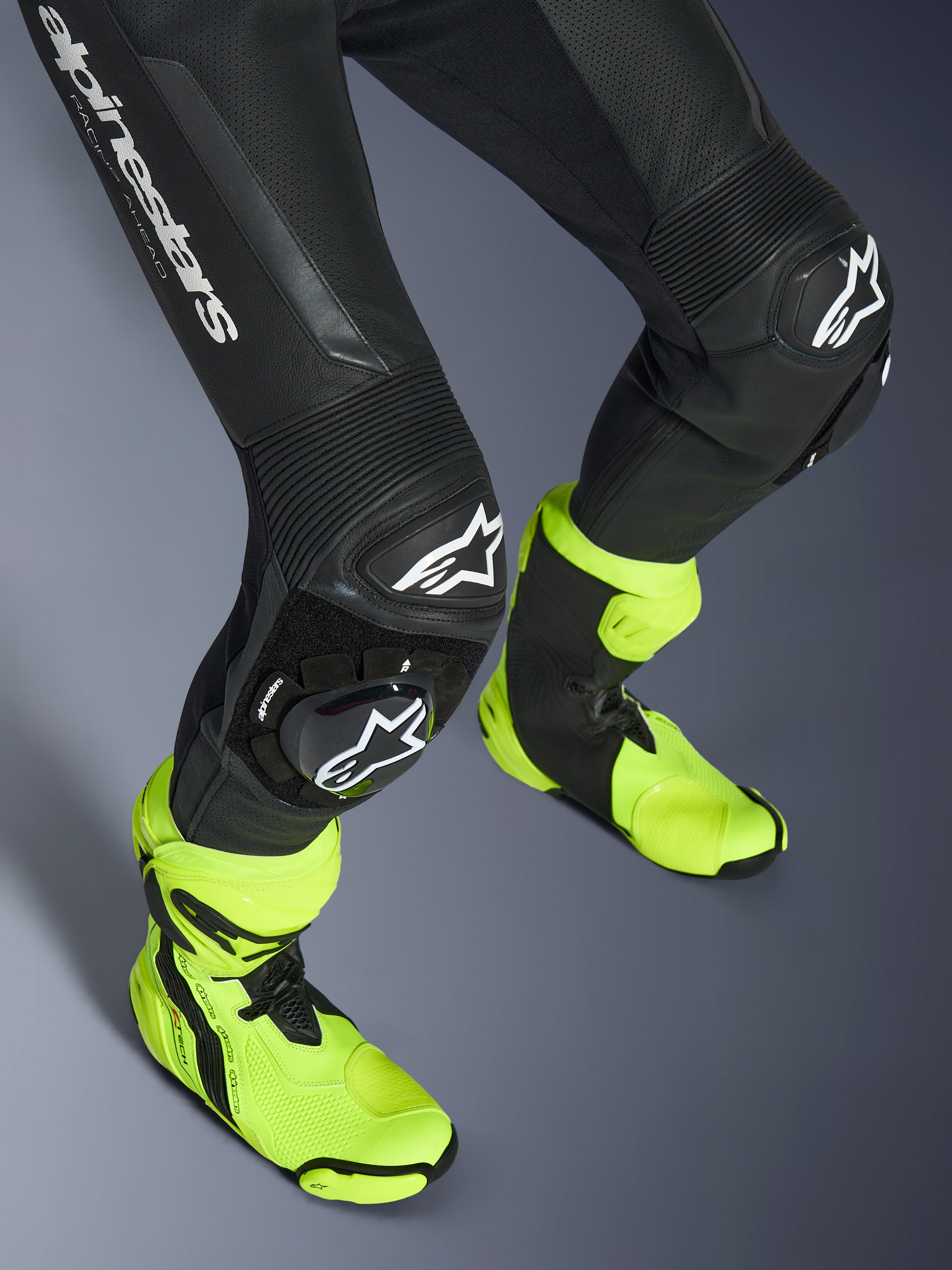 Combinaison en cuir Alpinestars GP Force V2, combinaison une pièce en cuir noir, gros plan sur les jambes et les genoux avec sliders à logo blanc, panneaux extensibles en accordéon, portée avec des bottes de course jaune fluo, équipement de course moto haute performance