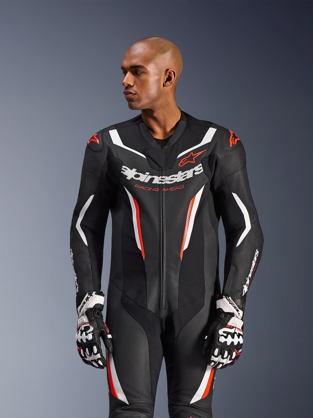 Combinaison en cuir Alpinestars GP Force V2 1 pièce, Noir Blanc Rouge Fluo, portée par un mannequin homme avec des gants de course, dotée d'une construction en cuir perforé, d'accents graphiques blancs et rouges, et du logo Alpinestars sur la poitrine.