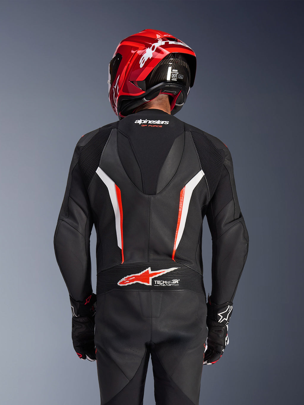 Combinaison en cuir Alpinestars GP Force V2 1 pièce, noir, blanc et rouge fluo, vue de dos d'un pilote portant un casque de course rouge et des gants, avec panneaux en cuir perforé, bosse aérodynamique avec logo Alpinestars et logo Tech-Air Ready dans le bas du dos, équipement de course moto professionnel.